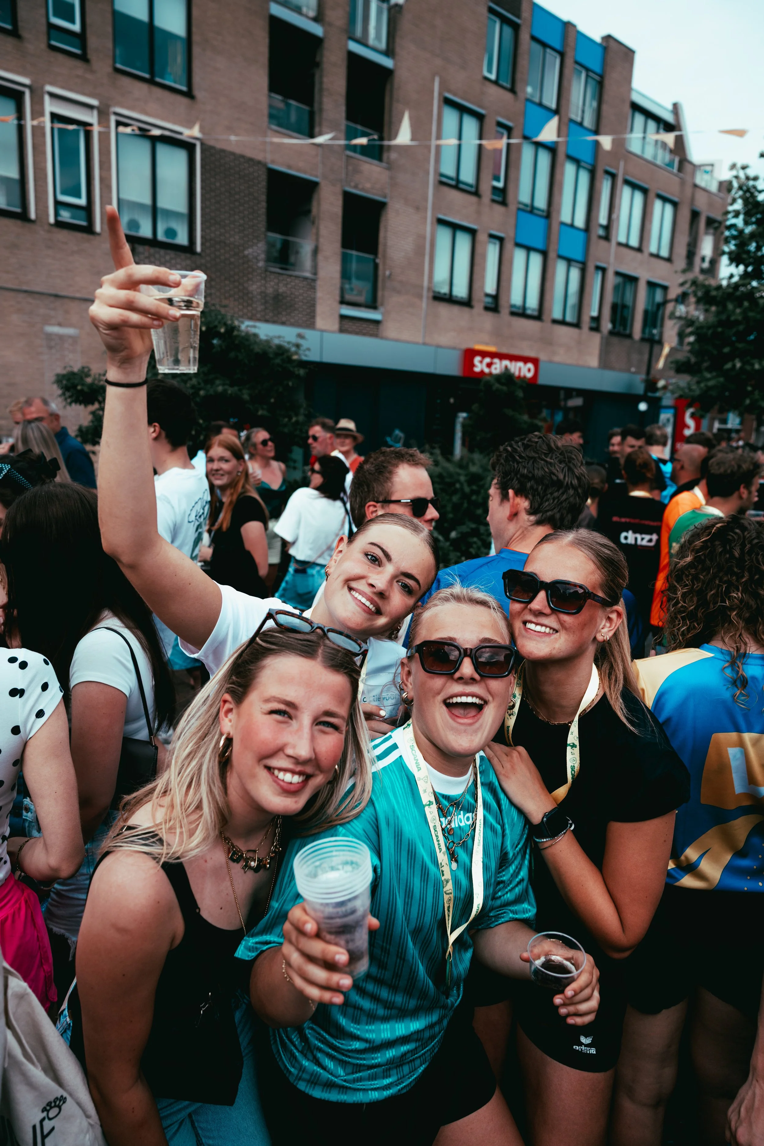 A group of people smiling and holding drinks at an outdoor event with a crowd and buildings in the background.