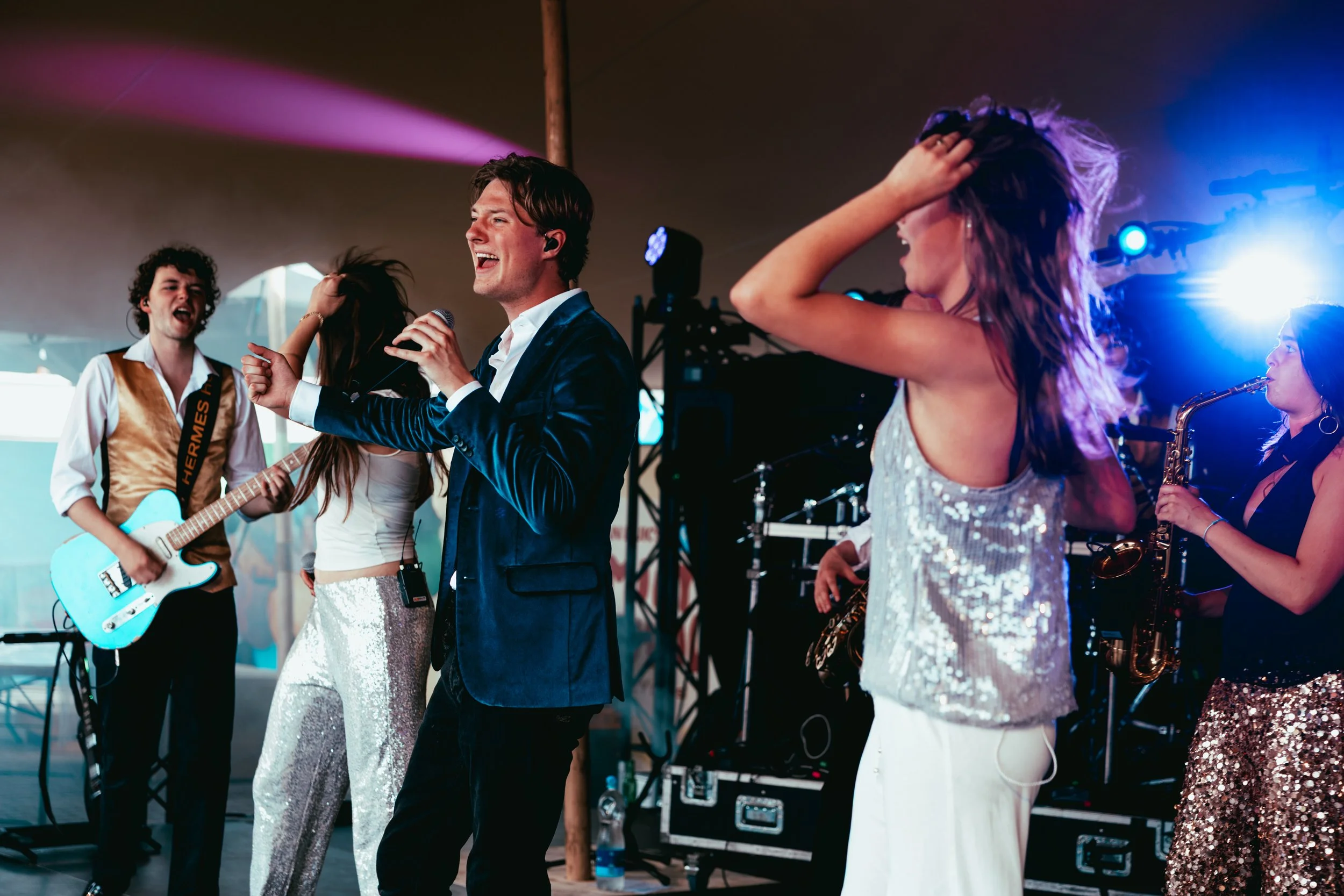 A group of musicians and singers performing on stage under colorful lighting, with one person holding a guitar and others singing and playing instruments.
