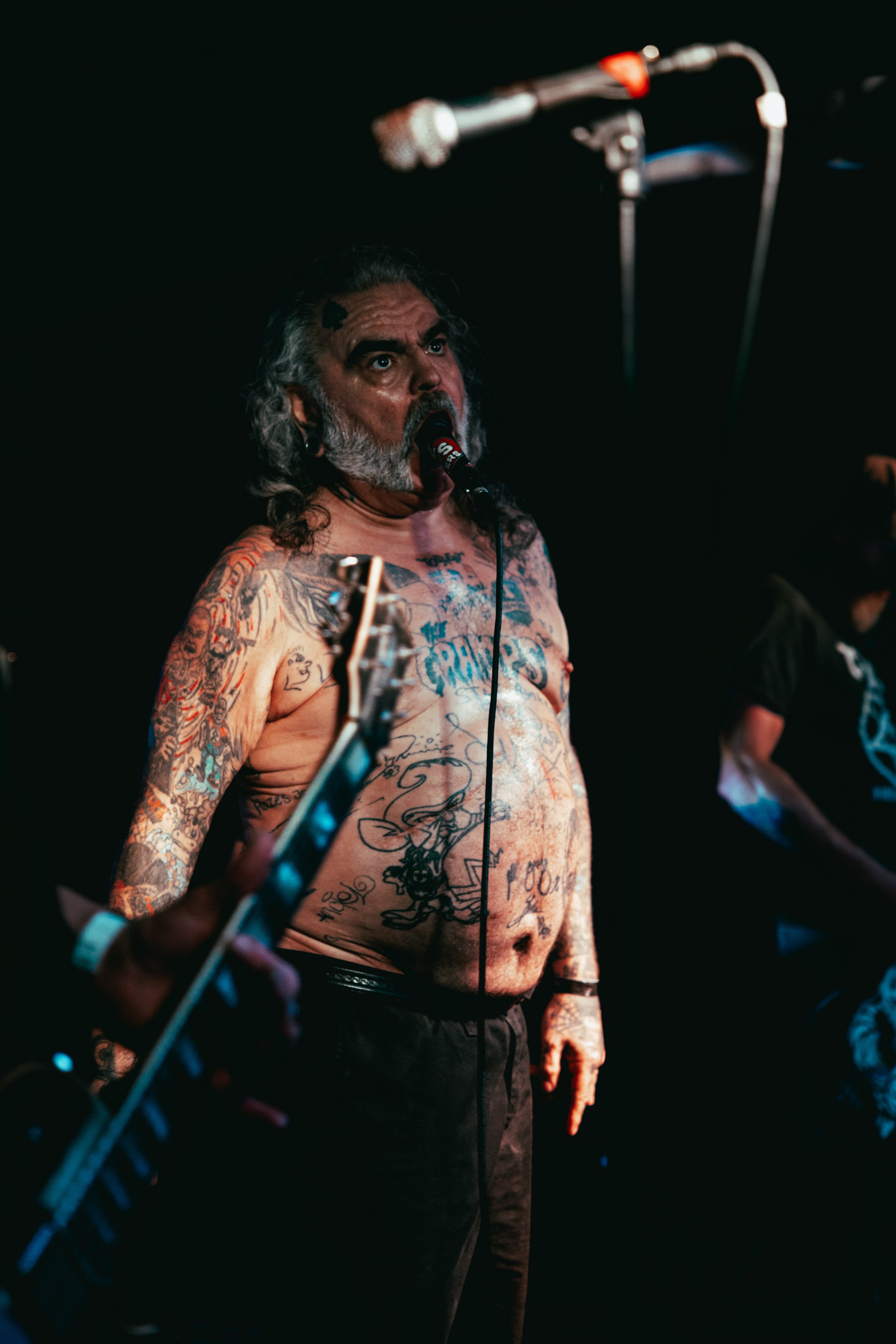 Man with long gray hair and beard singing into a microphone, shirtless with numerous tattoos, in a dark setting.