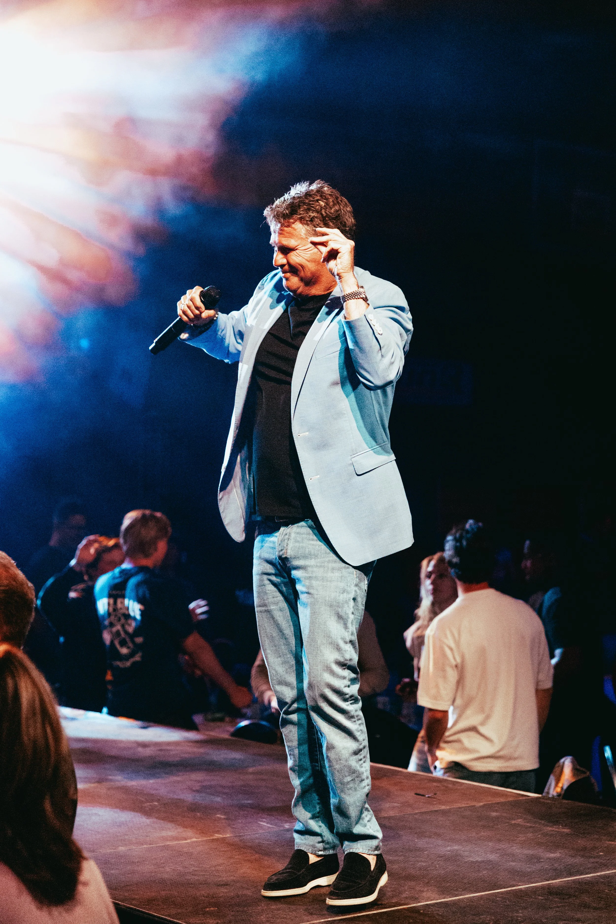 A man in a light blue blazer and jeans holding a microphone on stage, speaking to an audience with a dark background and colorful stage lighting.