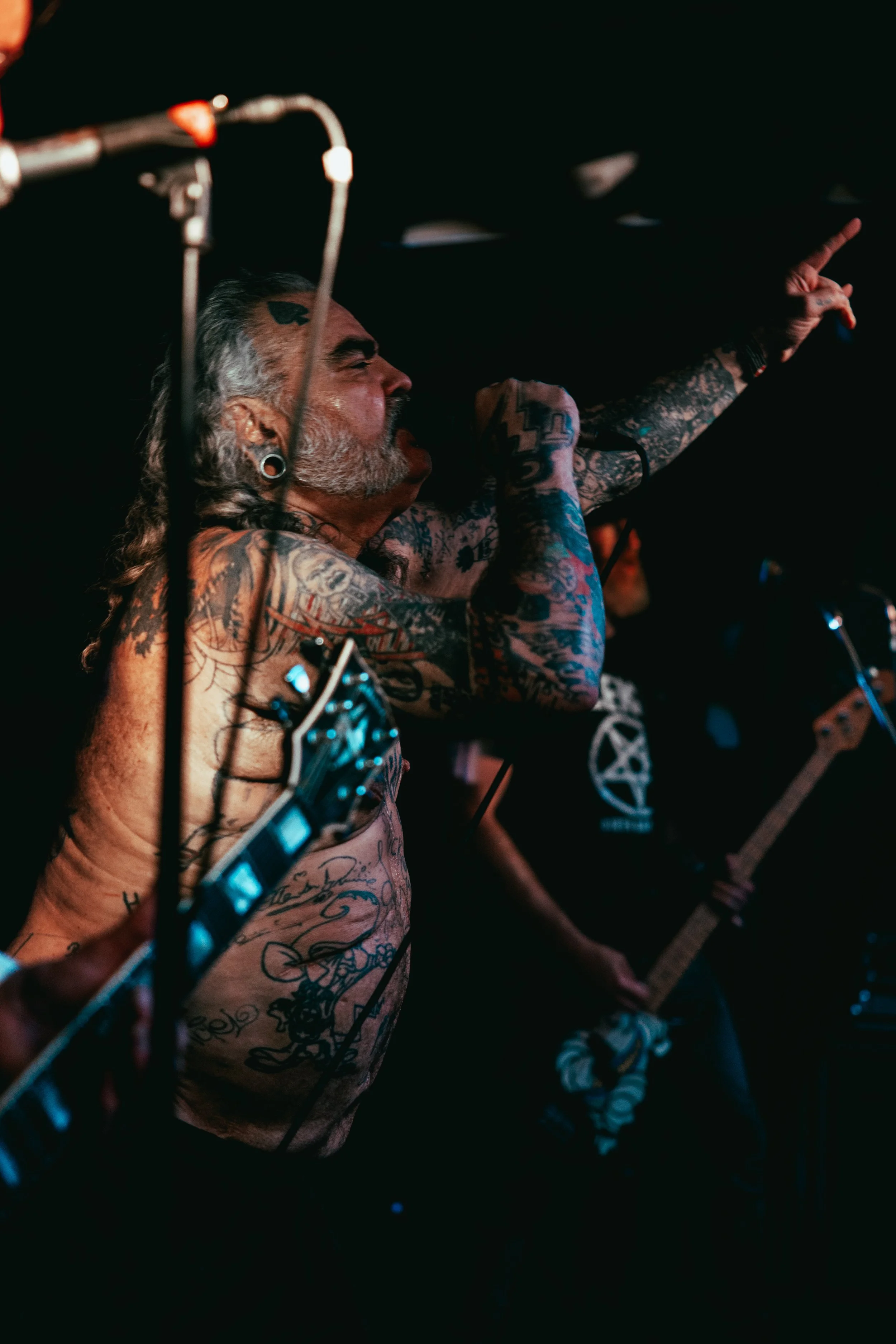 A shirtless man with tattoos and a beard playing an electric guitar on stage, pointing upwards with his right hand, in a dark setting.