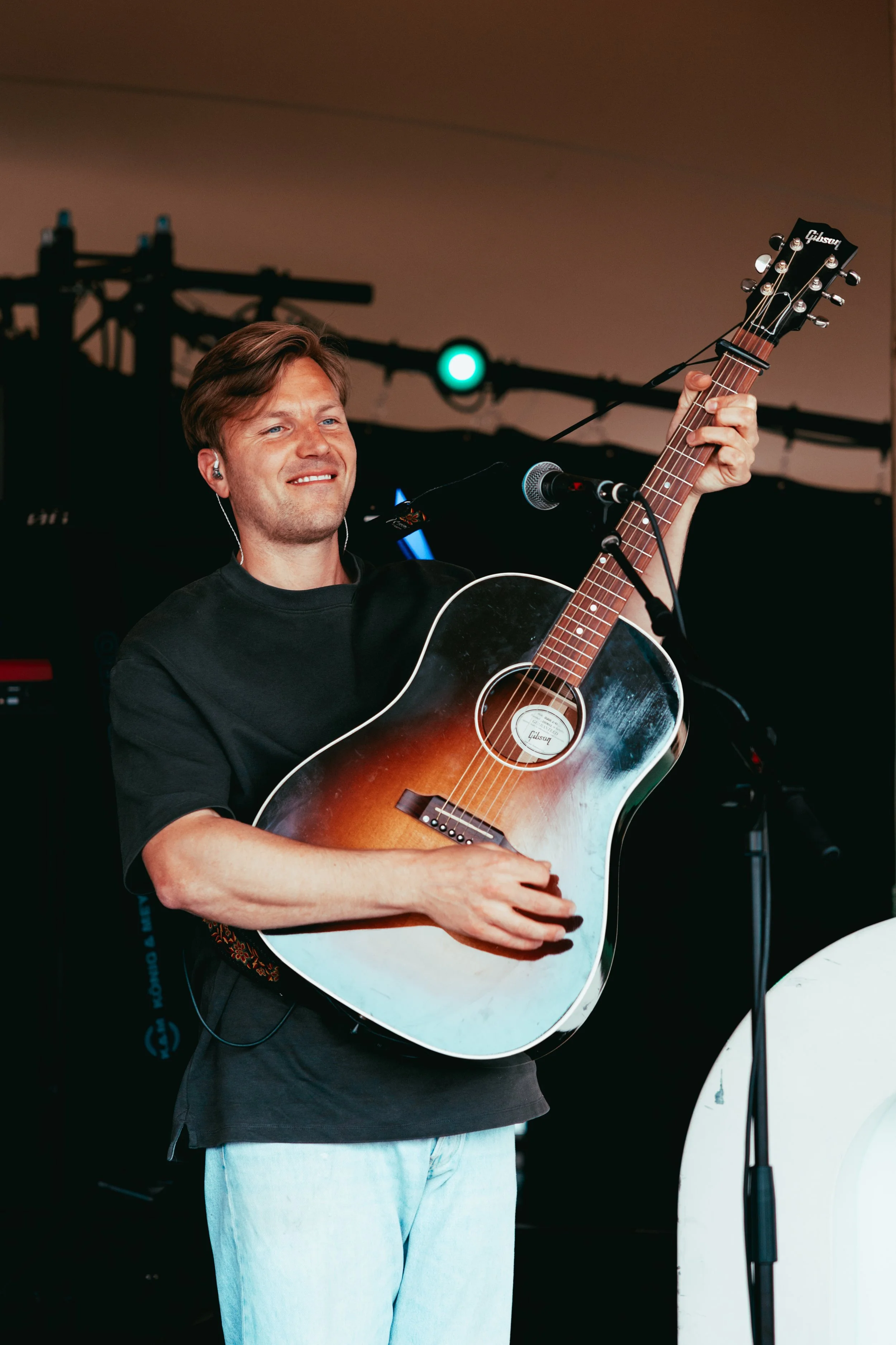 A young man with brown hair and light skin playing an acoustic guitar on stage. He is smiling and wearing earbuds, with a microphone in front of him. The background has stage equipment and a green stage light.