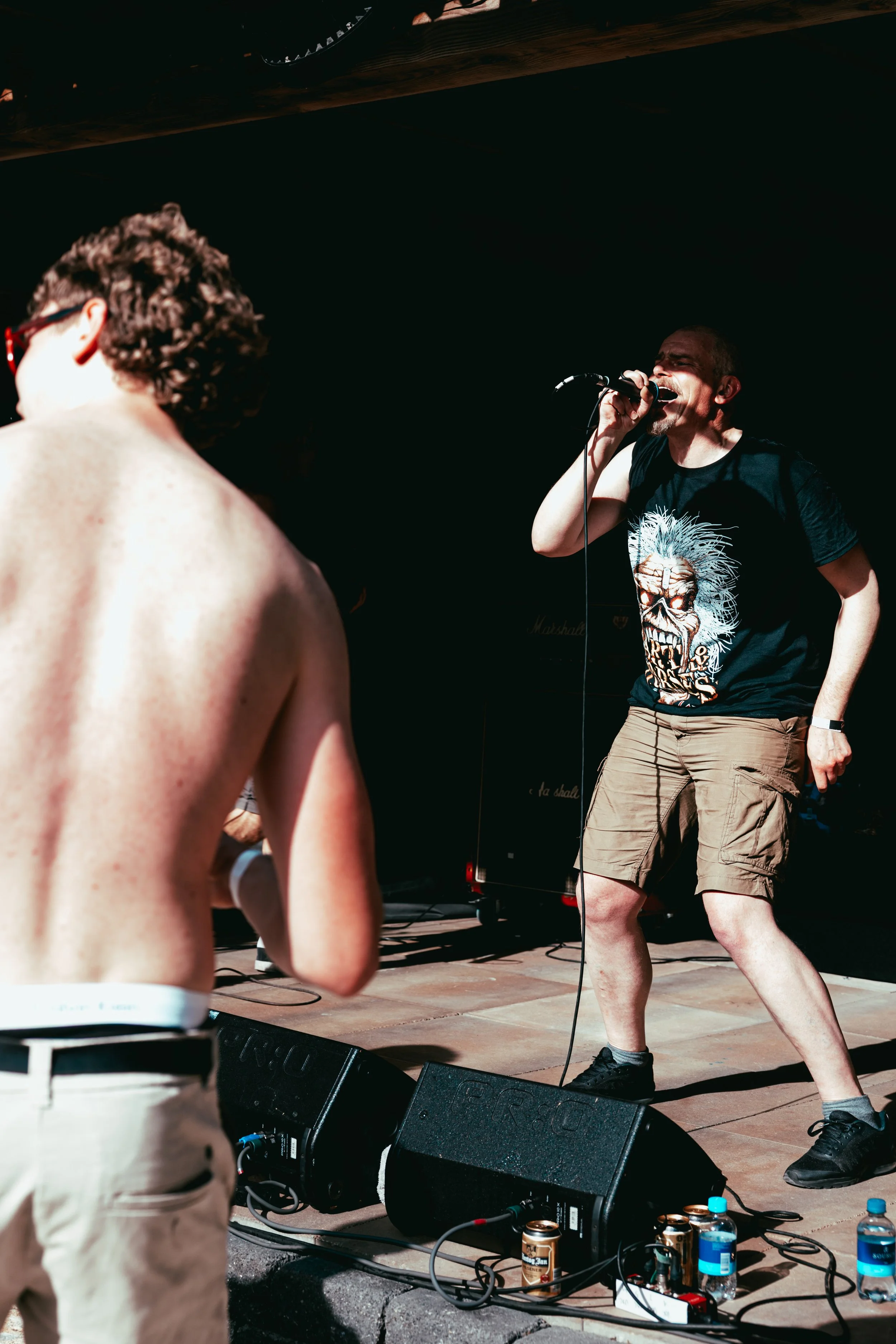 A vocalist passionately singing into a microphone during a live outdoor music performance, with a guitarist slightly blurred in the foreground and musical equipment including speakers and water bottles on the stage.