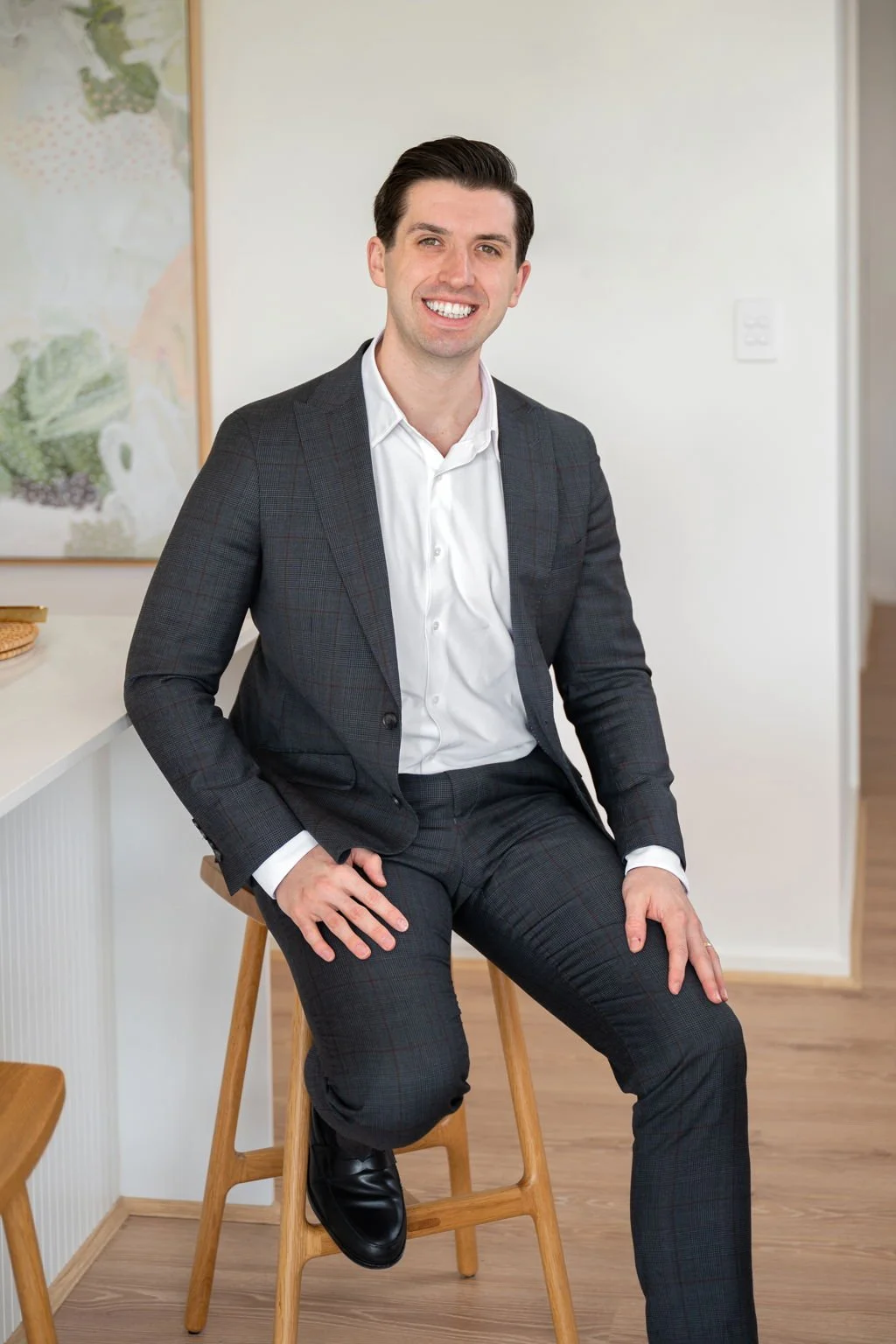 A smiling man in a dark checkered suit and white shirt sitting on a wooden stool in a modern room.