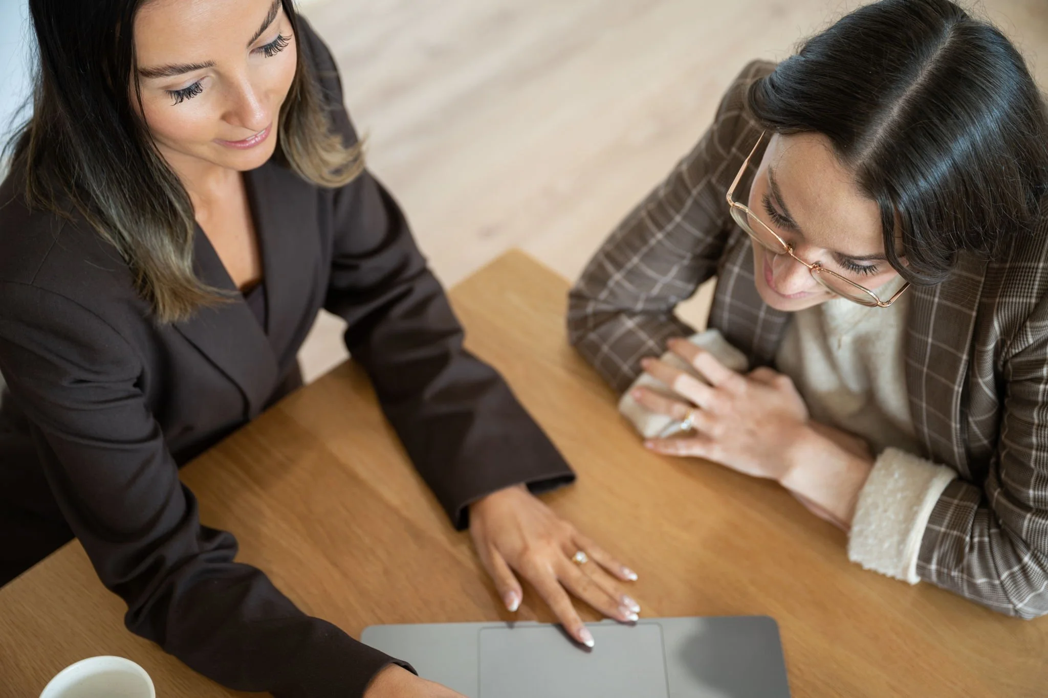 Two women are seated at a wooden table, looking at a laptop. One woman is wearing a black blazer, and the other is wearing glasses and a plaid blazer. They appear focused and engaged in a discussion or work.