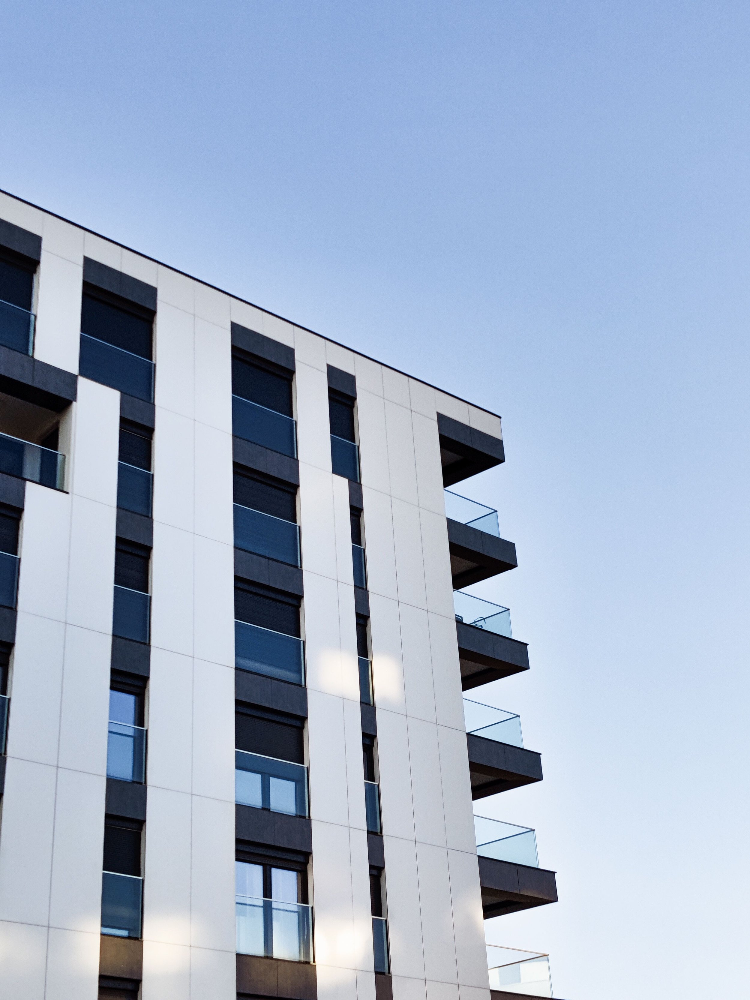 Modern multi-story building with white and dark gray exterior panels, large glass windows, and balconies against a light blue sky.