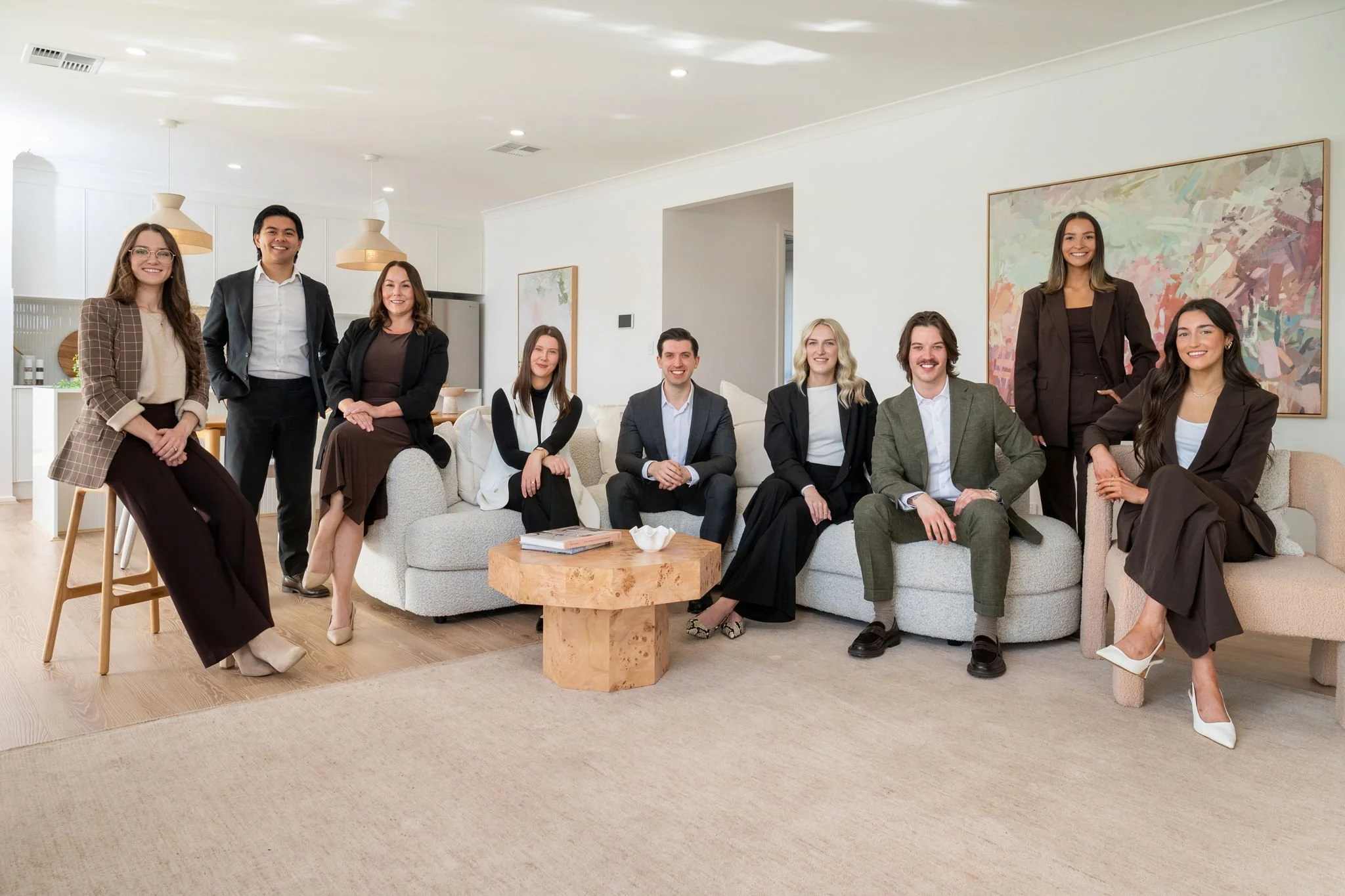 Group of ten diverse professionals in business attire posing in a modern living room with a beige sofa, wooden coffee table, and abstract artwork on the wall.