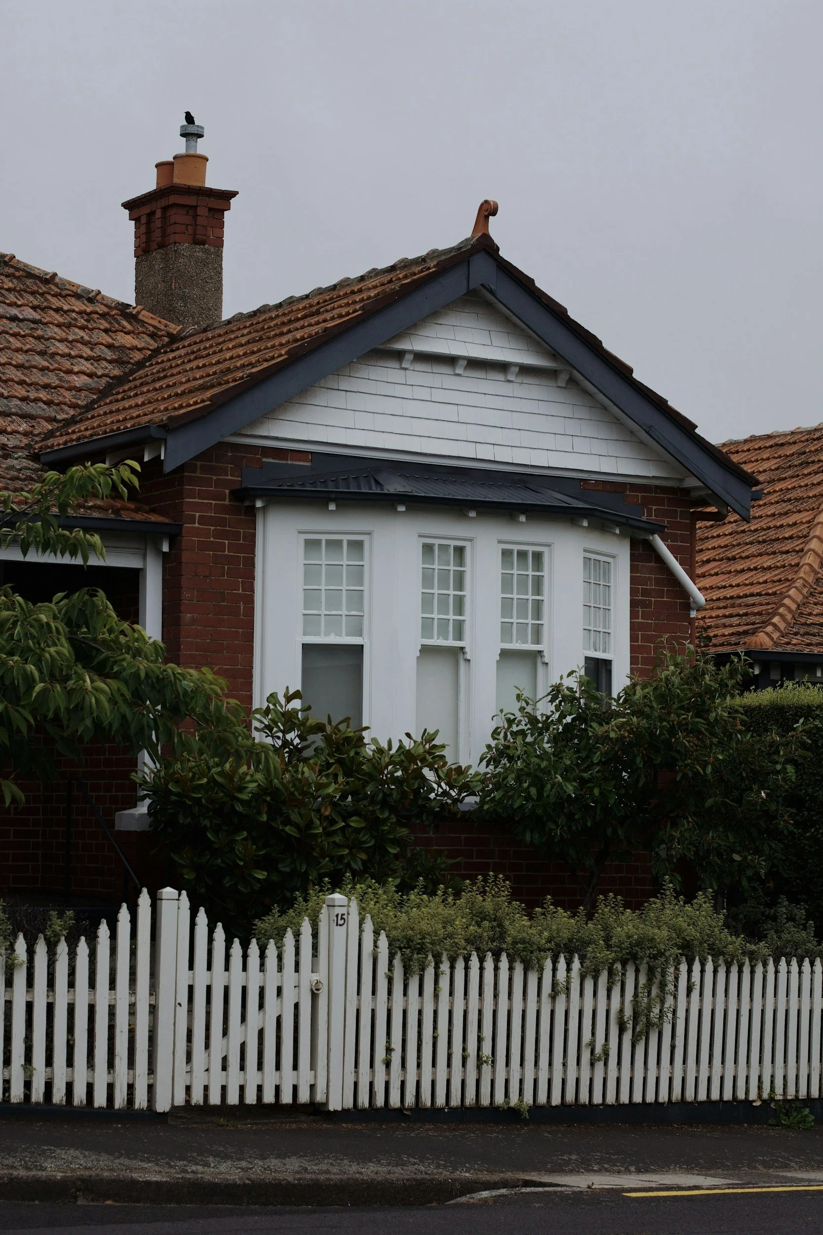 A house with a brick facade, white windowed bay, and a white picket fence, surrounded by greenery. The roof has red tiles and a chimney with a bird on top, and the sky is overcast.