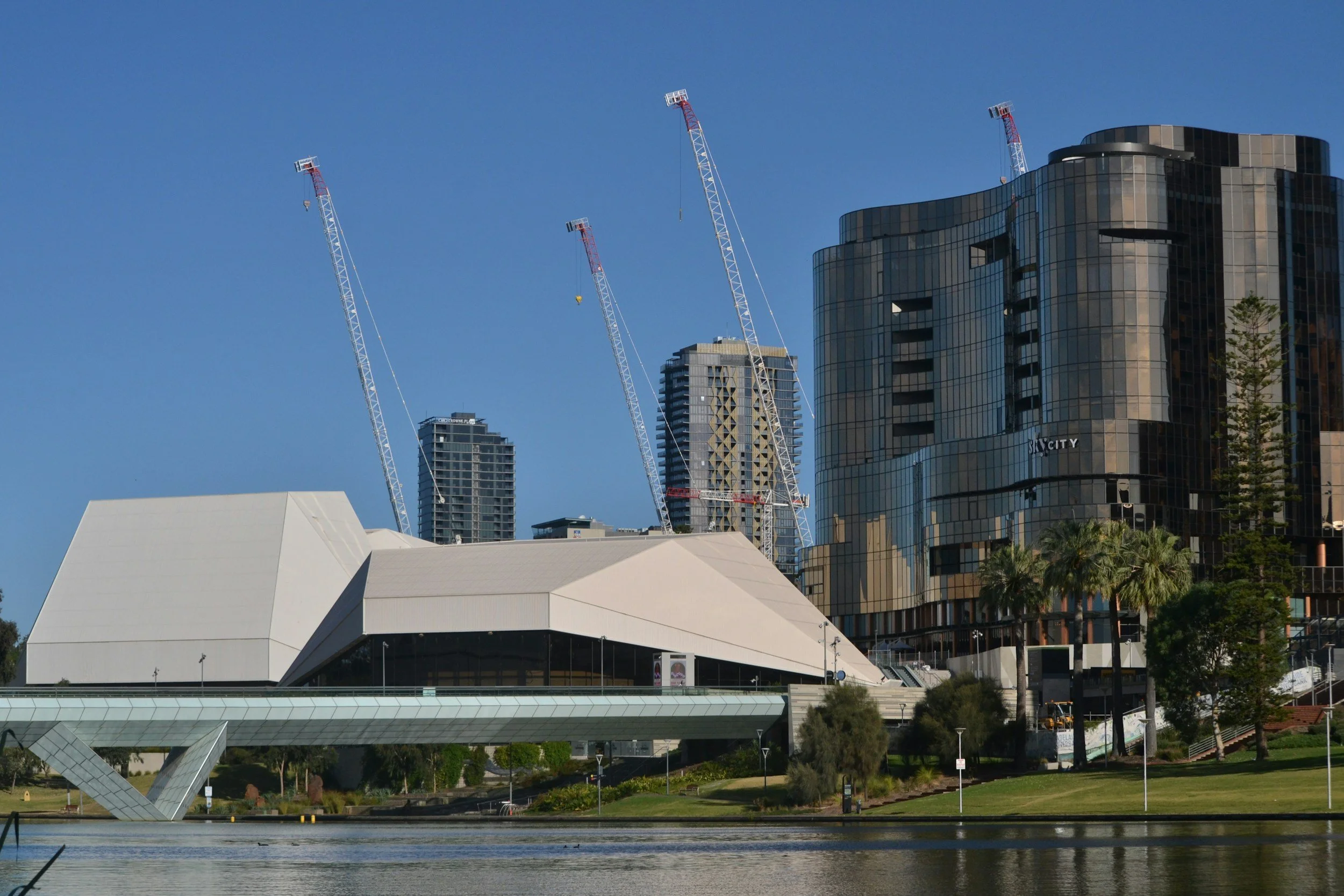 High-rise buildings with glass exteriors, construction cranes, a modern white building with angular design, and a body of water with trees and greenery in the foreground.