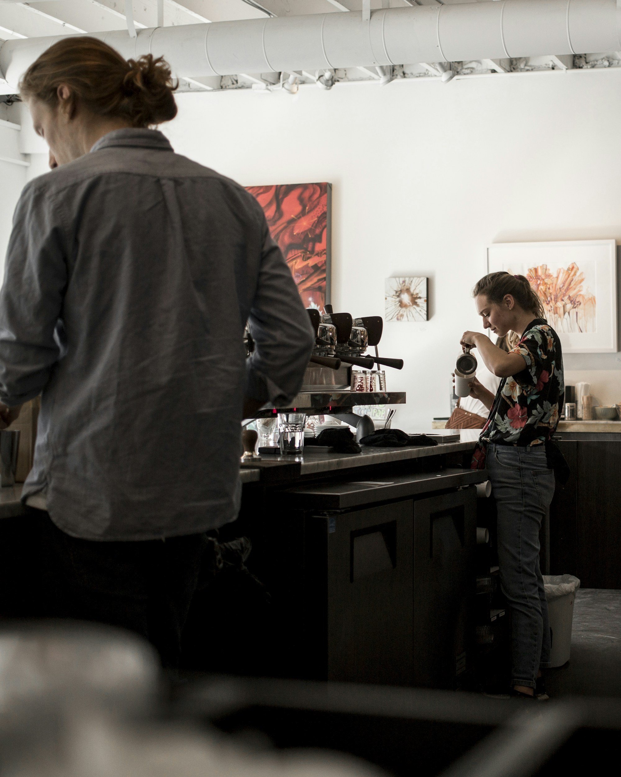 A woman in a floral shirt preparing to use a microscope at a barista counter in an art gallery with paintings on the wall.