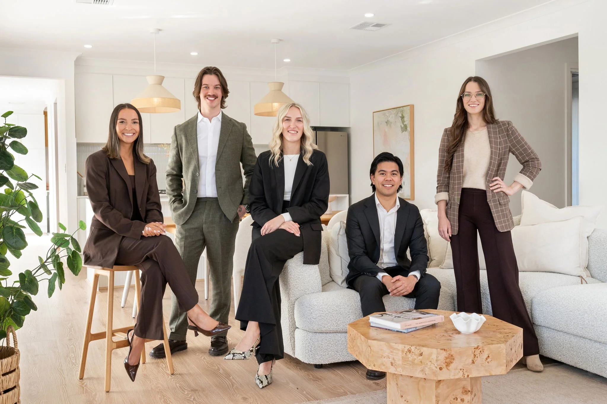 Group of six professionals in an office living room, smiling at the camera. Two women are seated, one standing on the left, one on the right. Two men and one woman are standing behind them. Bright, modern space with white walls, wood accents, and a large green plant.