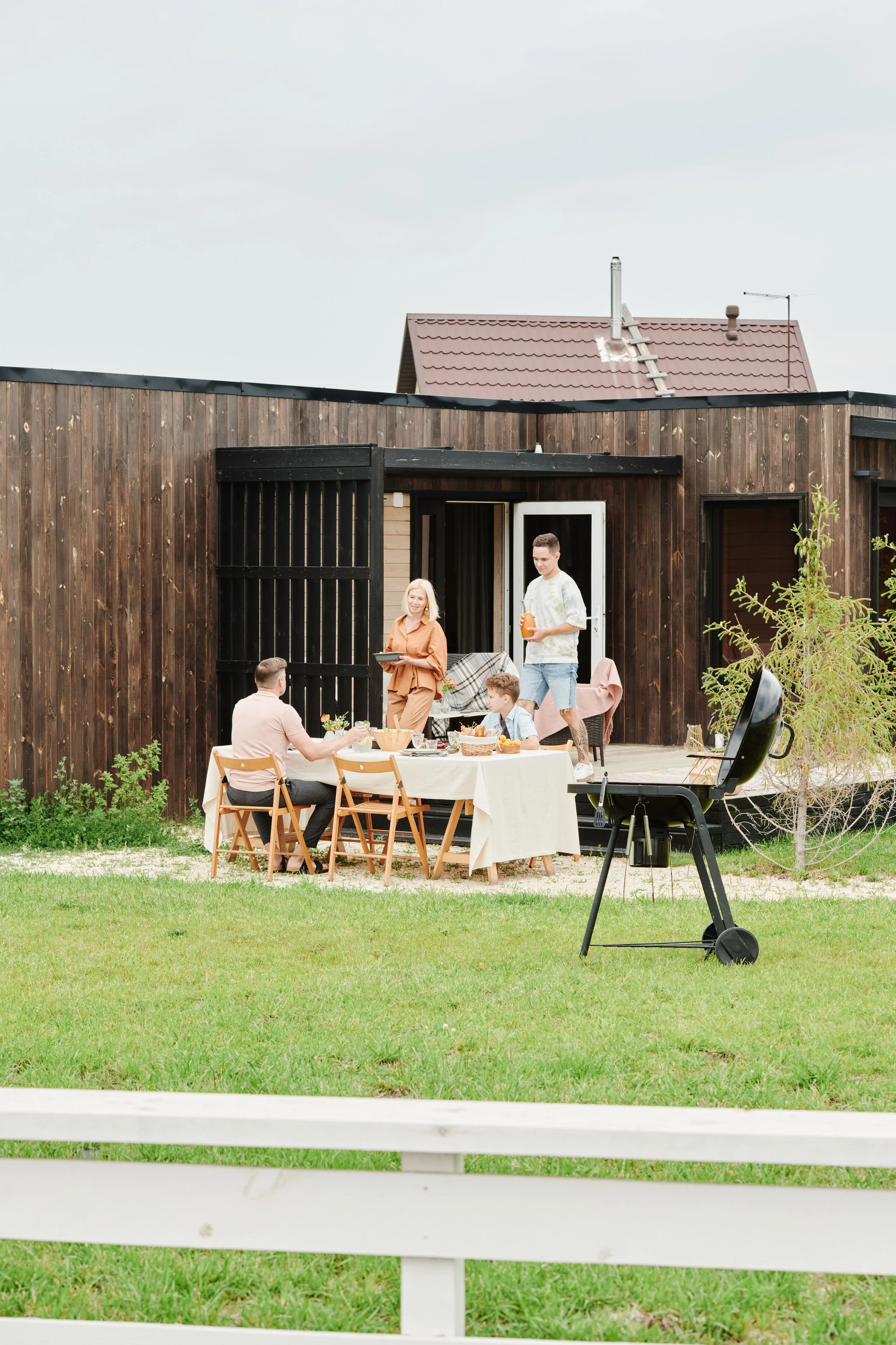People having a backyard barbecue or outdoor gathering in front of a wooden house with a red roof, a chimney, and outdoor furniture, with a black grill in the foreground.