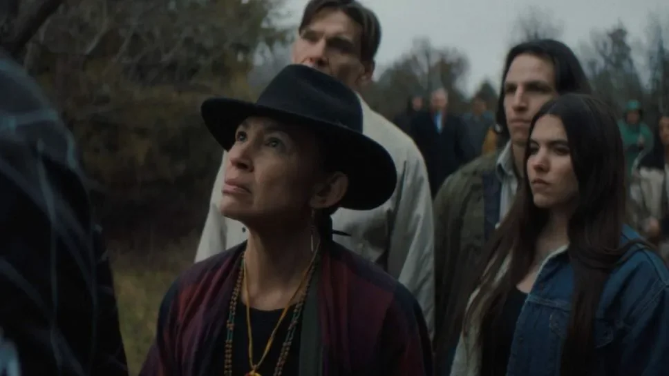 A group of people, including a woman wearing a black hat and layered necklaces, stand outdoors with serious expressions during daytime. The background shows trees and a cloudy sky.