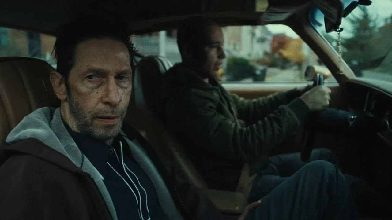 Two men sitting in the front seats of a car, the man in the passenger seat has a disheveled appearance with rough skin and looks directly at the camera, while the man in the driver's seat is focused on driving, with an urban neighborhood visible through the windshield.
