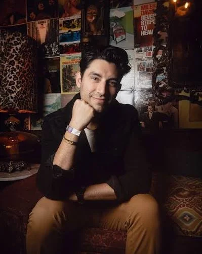 A young man with dark hair and a light beard sitting on a colorful patterned sofa in a dimly lit room. He's resting his chin on his right hand and smiling at the camera, with a background of posters and artwork on the wall.