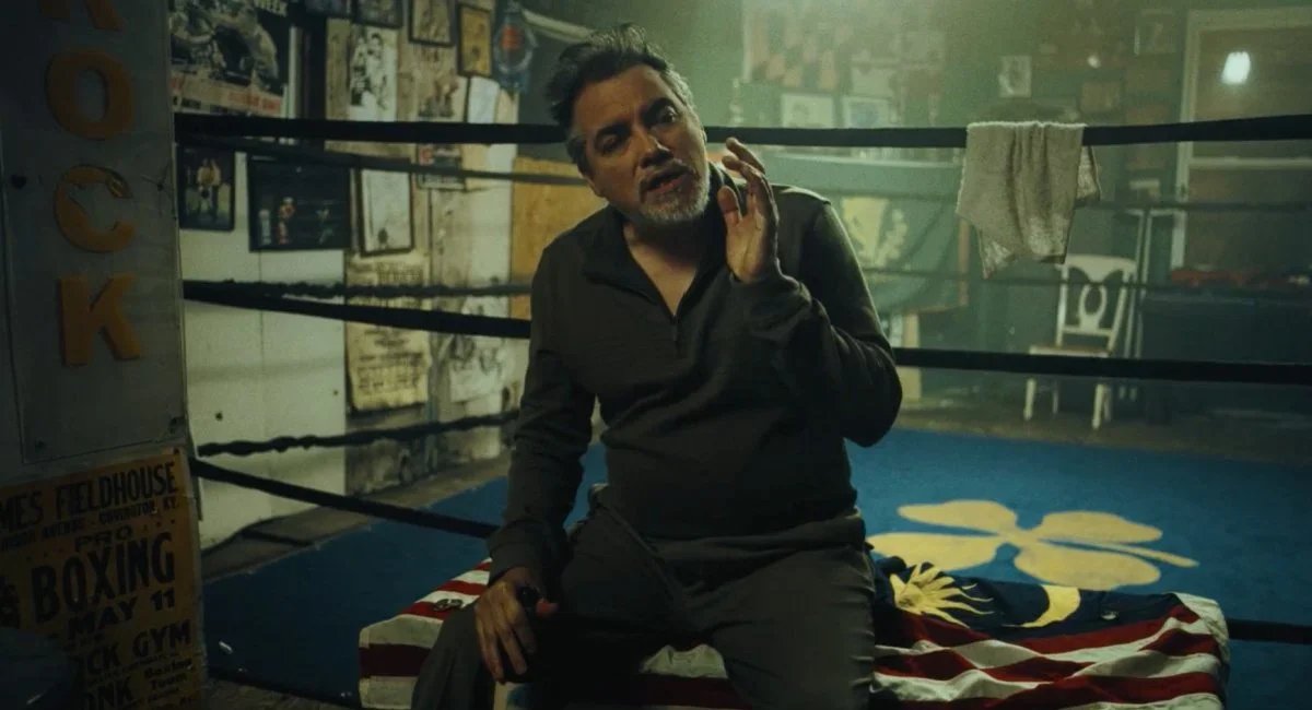 A man sitting inside a boxing gym with boxing rings, posters, and equipment in the background, speaking or singing with his hand near his face.