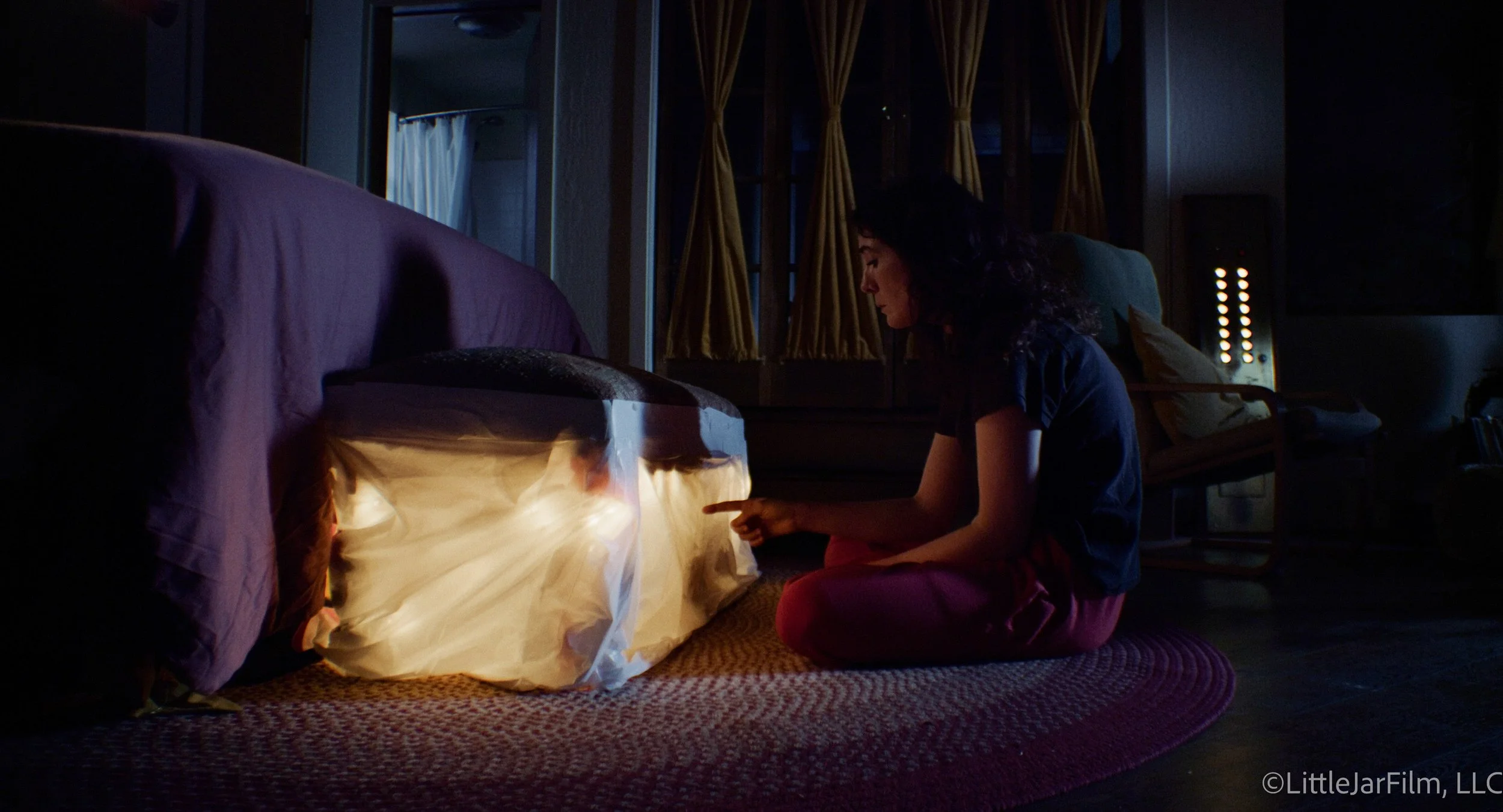 A woman with dark, curly hair is sitting on a round, patterned rug in a dimly lit room, pointing at a glowing, illuminated fabric or paper in front of her. She is wearing a dark top and red pants, and appears to be focused on the illuminated object.