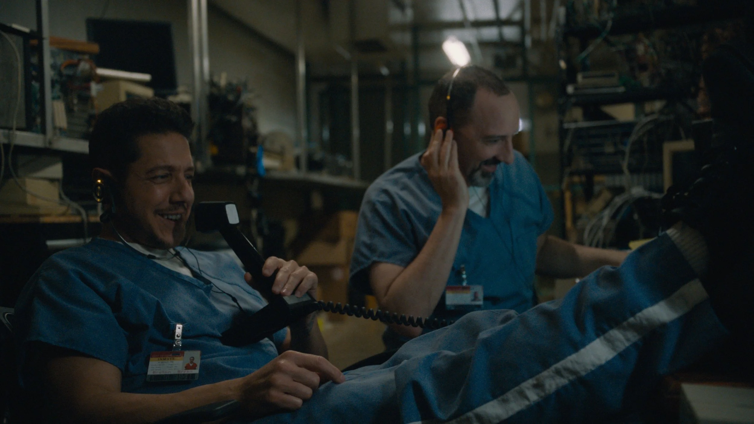 Two astronauts in blue uniforms relaxing in a spacecraft or space station, smiling and talking, with various equipment and shelving in the background.