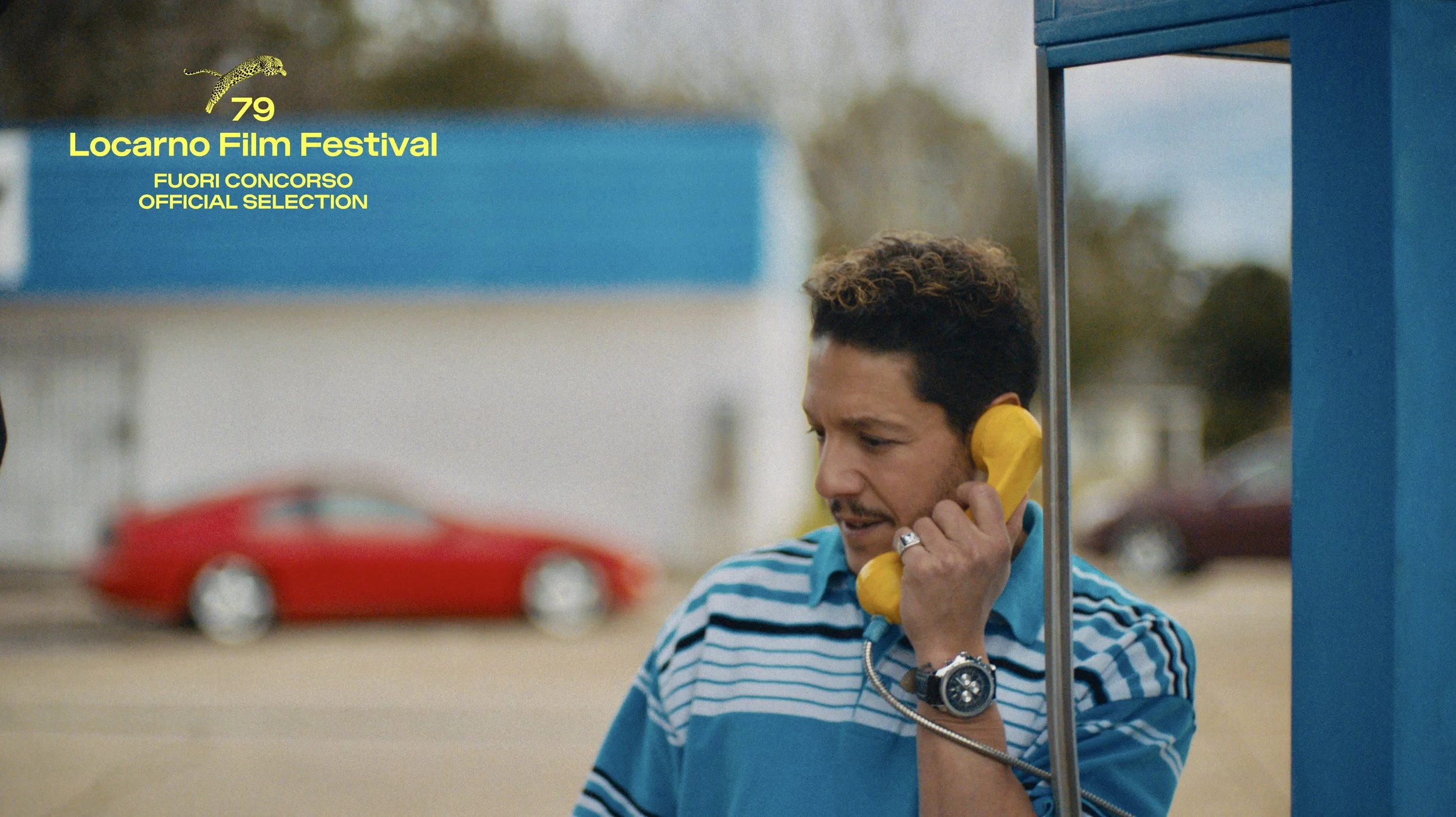 A man with curly hair and a beard, wearing a striped blue shirt, talking on a yellow phone at a phone booth.