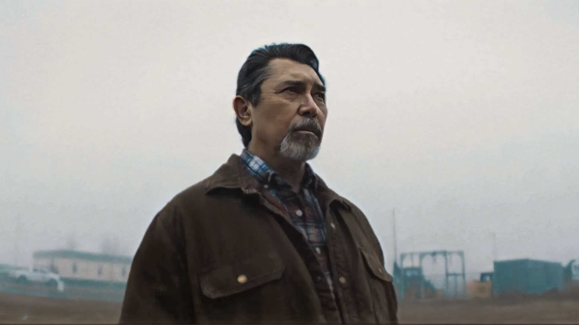 A man with dark hair and a beard standing outdoors near a foggy area with buses and buildings in the background.