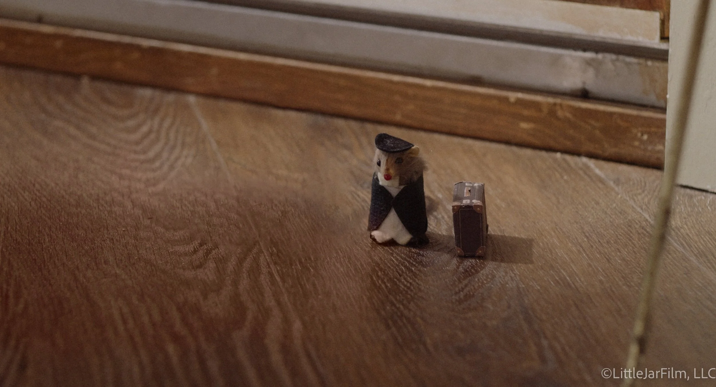 A small toy mouse dressed as a traveler with a black hat and coat, standing on a wooden floor next to a miniature suitcase.