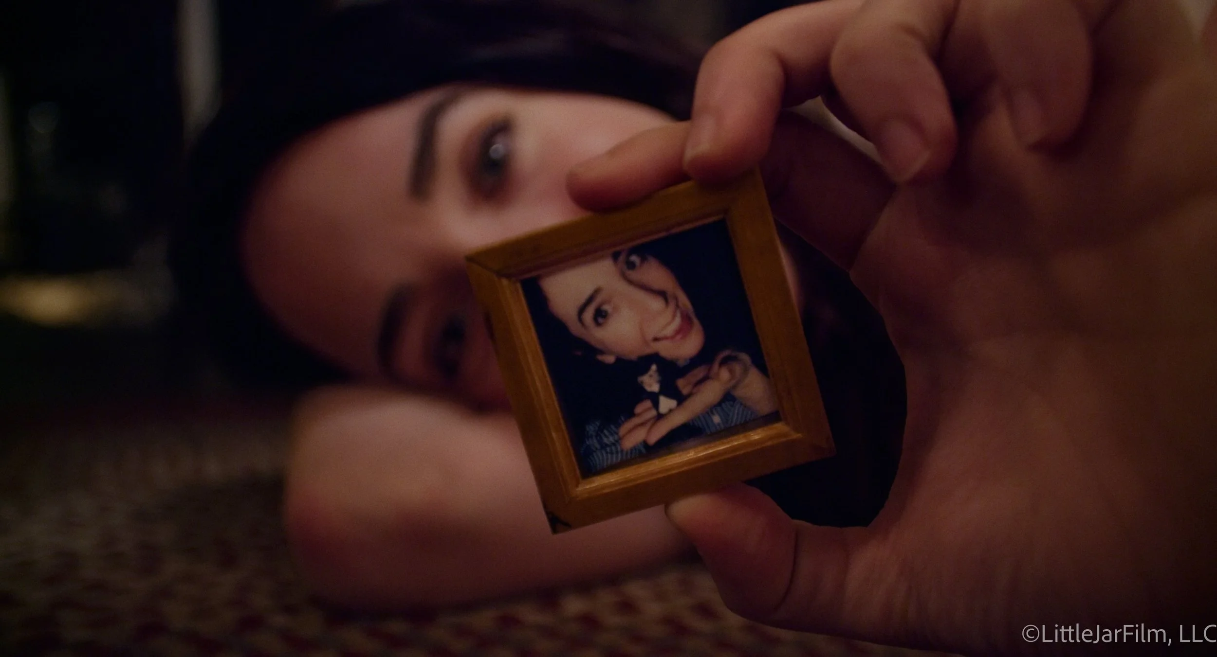 Close-up of a person lying down, holding a small framed photograph of a smiling woman with dark hair, wearing a checkered shirt.