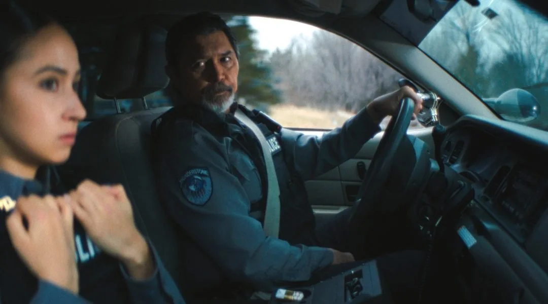 Two police officers sitting inside a police vehicle, looking serious, with the officer in the driver's seat looking toward the camera and the other officer looking away.