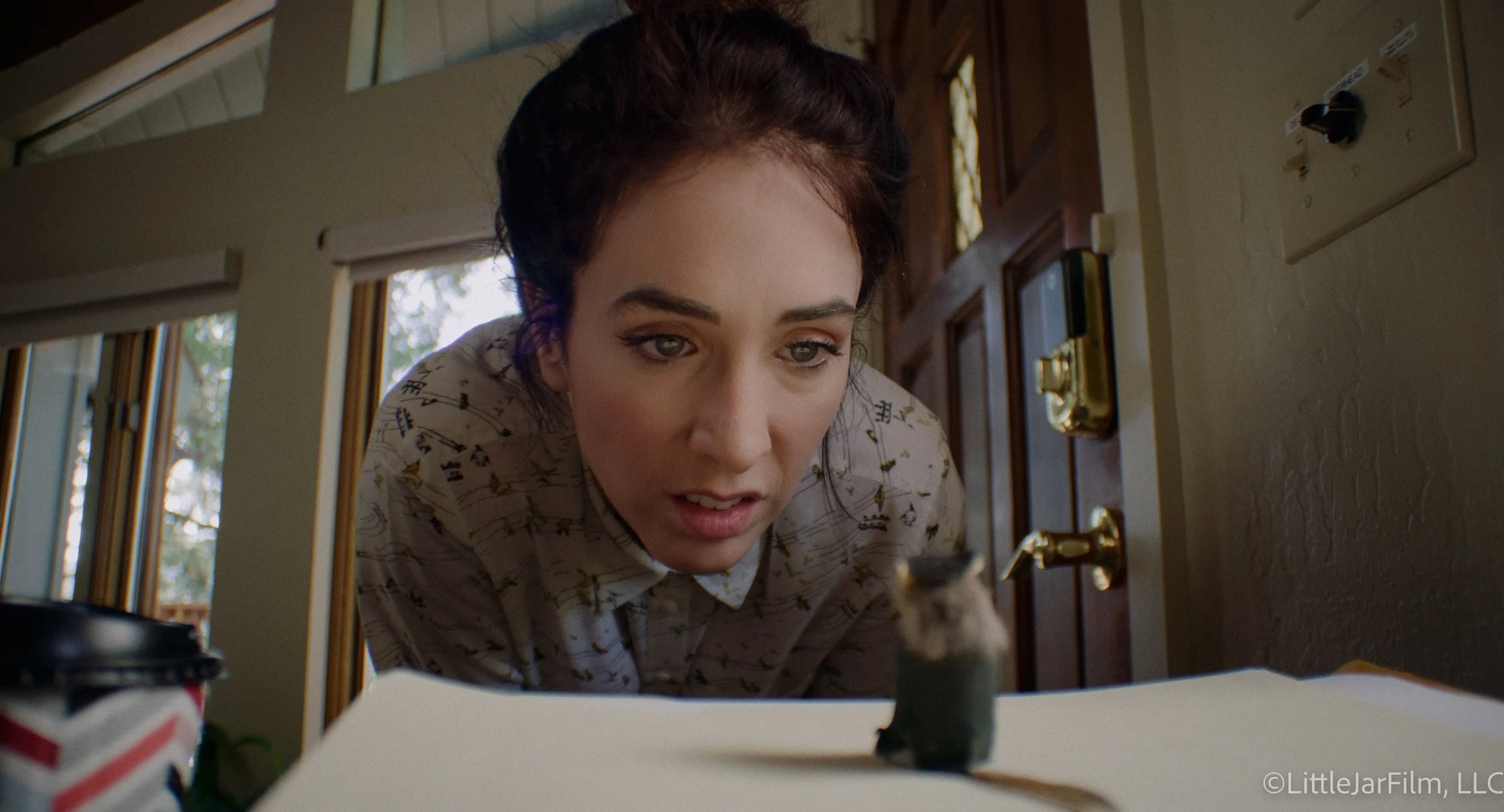 A woman with dark hair and light skin leaning over a surface, looking at a small animal, possibly a kitten or a puppy. The background shows a wooden door and windows with curtains, indicating an indoor setting.