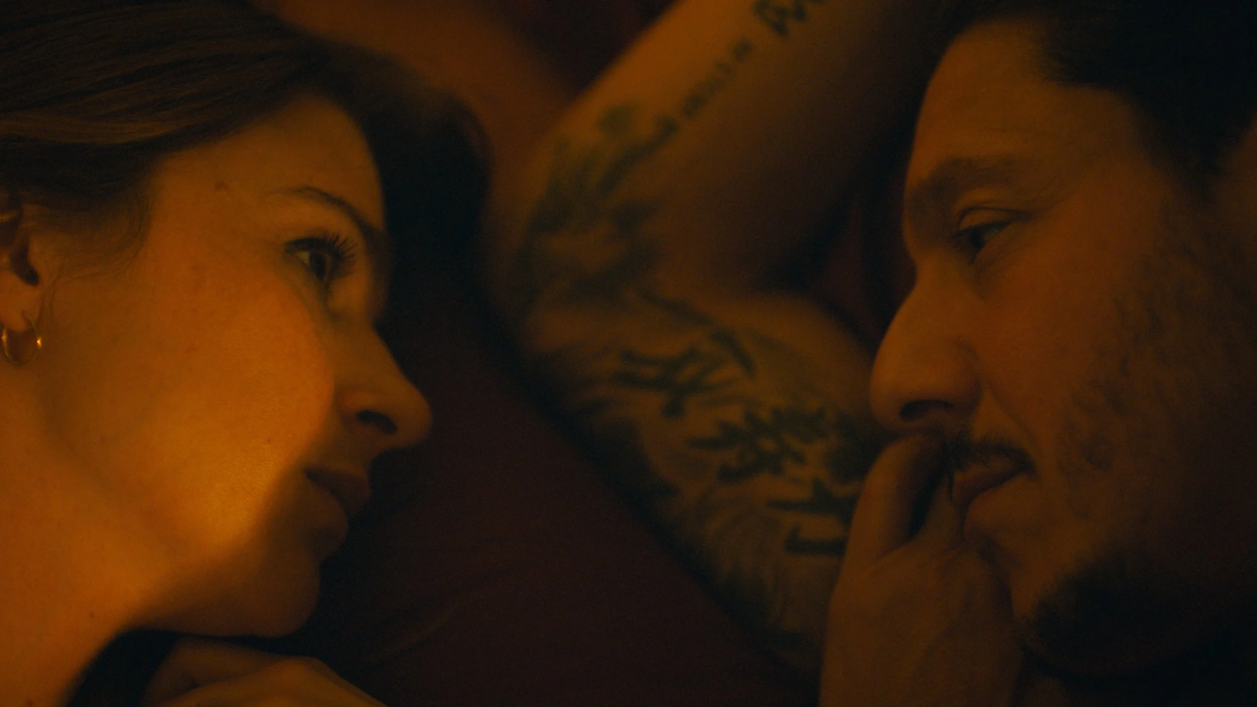 Close-up of a man and a woman lying face to face with their foreheads touching, in a dimly lit setting.