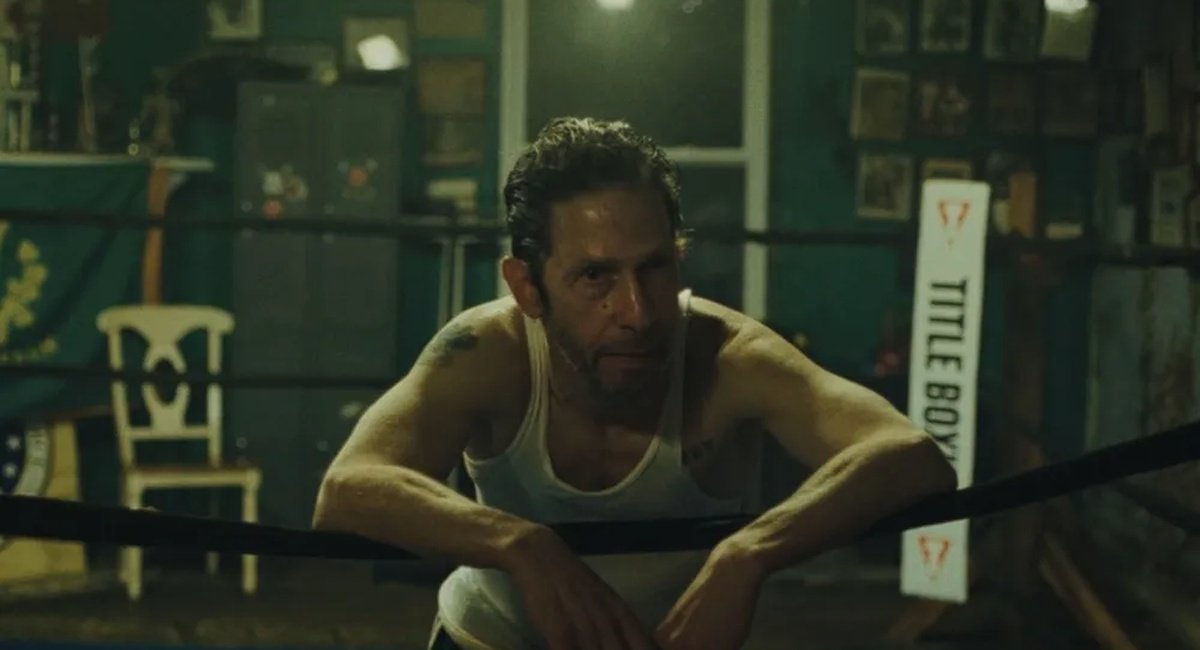 A man with a muscular build, wearing a white tank top, leaning on a boxing ring's ropes in a dimly lit gym, with boxing equipment and framed photos on the walls in the background.