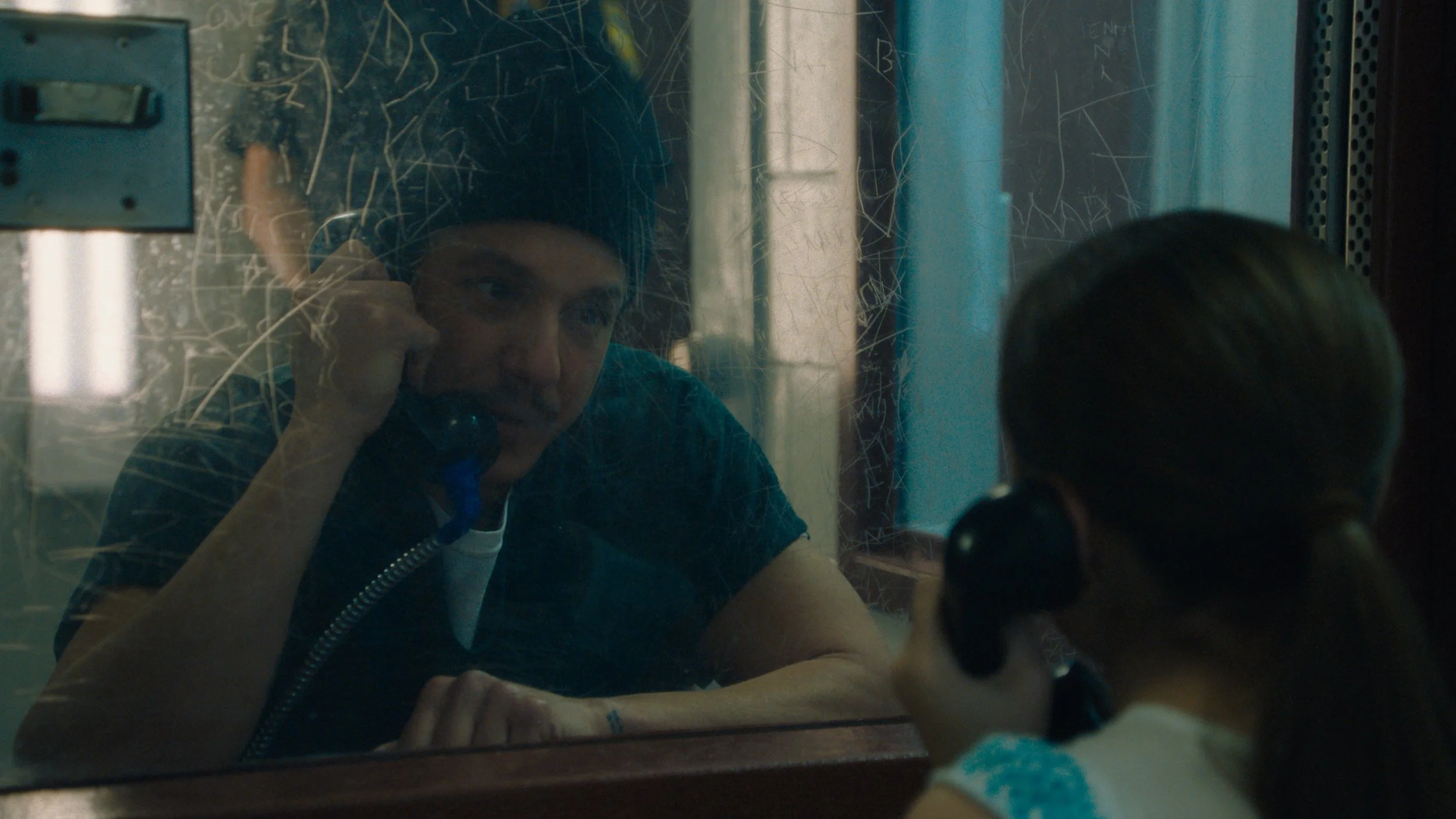 A person with dark hair speaking on the phone, seen through a scratched or dirty glass window, with another person visible from behind, indicating a conversation at a police station or similar setting.