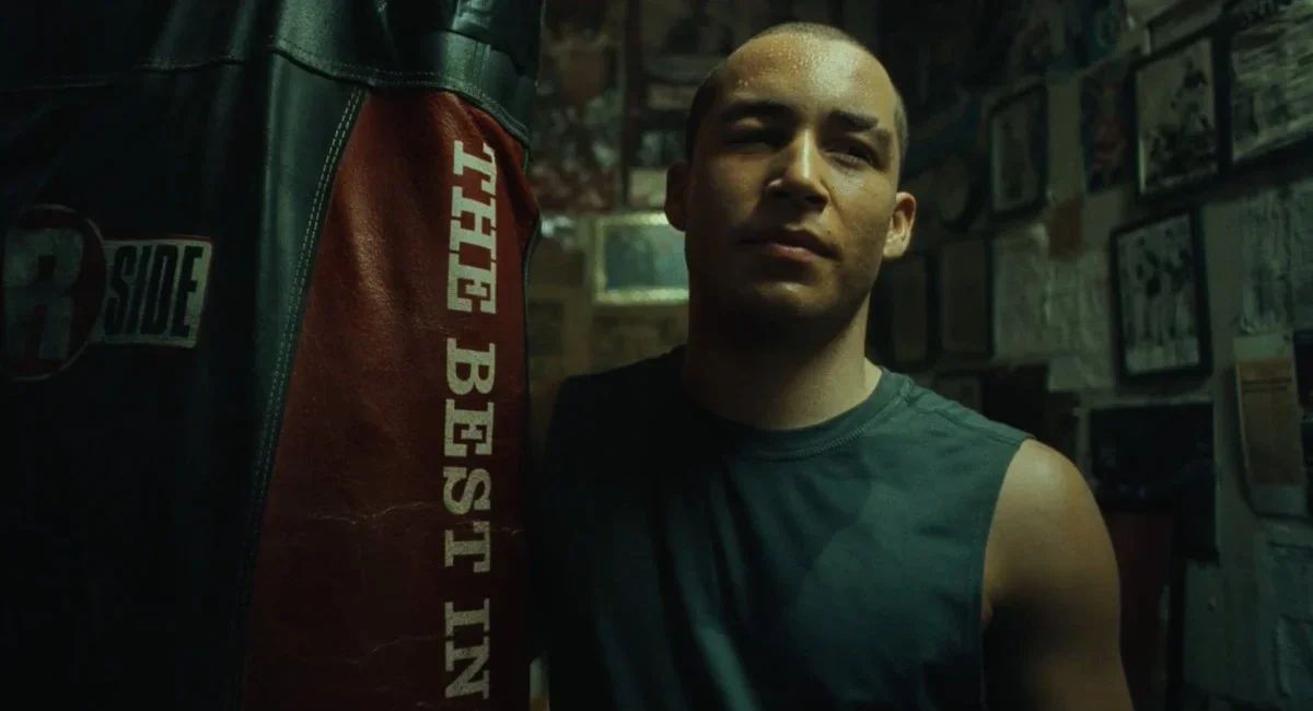 A close-up of a man with a shaved head wearing a sleeveless shirt, standing next to a punching bag in a gym or boxing gym with framed pictures on the wall.