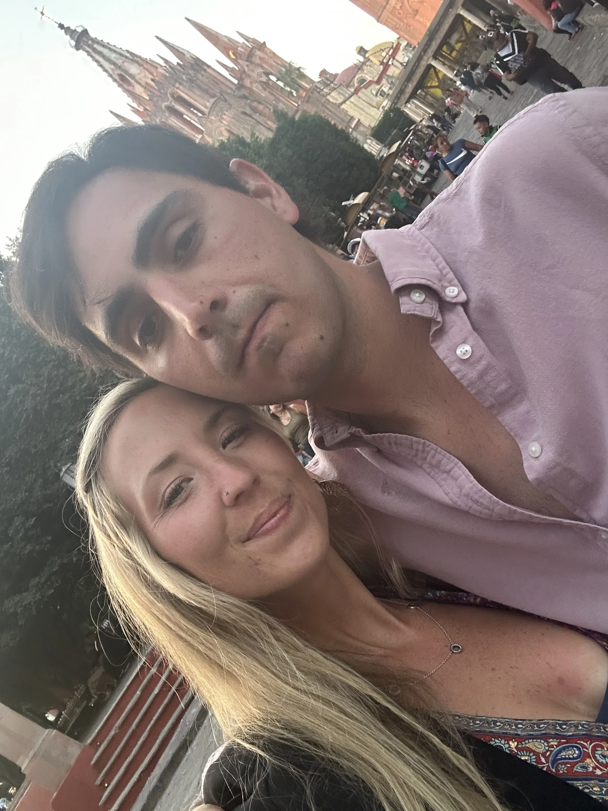 A selfie of a young man and woman outdoors in front of a historic cathedral with tall spires and a red brick facade. The woman has long blonde hair and is wearing a black top with a necklace, smiling at the camera.