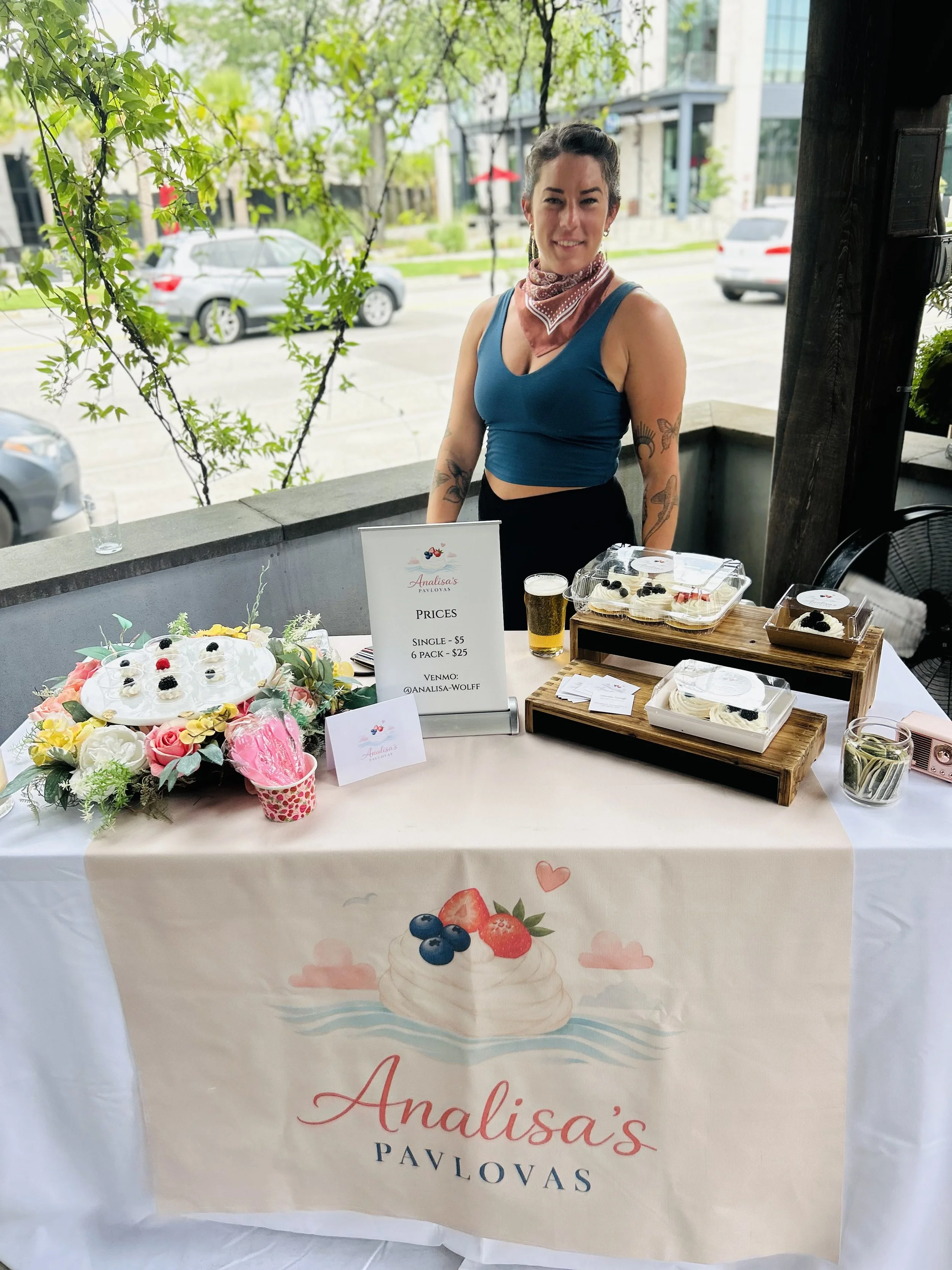 A woman standing behind a table with baked goods at an outdoor market stall. The table has a banner with the words "Analisa's Pavlovas" and a colorful illustration of pavlova with berries. There are cupcakes, a sign with prices, a glass of beer, and decorative flowers on the table. The woman is smiling, wearing a blue tank top, a bandana around her neck, and has tattoos on her arms. In the background, there are cars, buildings, and trees.