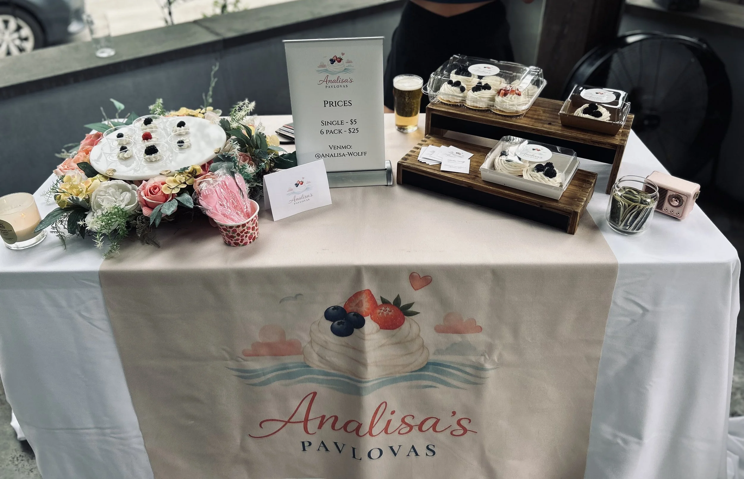 Table with a sign displaying prices for desserts from Analisa's Pavlovas, featuring a variety of pavlova desserts topped with berries, floral decoration, a pitcher of straws, a small pink camera, and business cards, with a background of vehicles and outdoor setting.