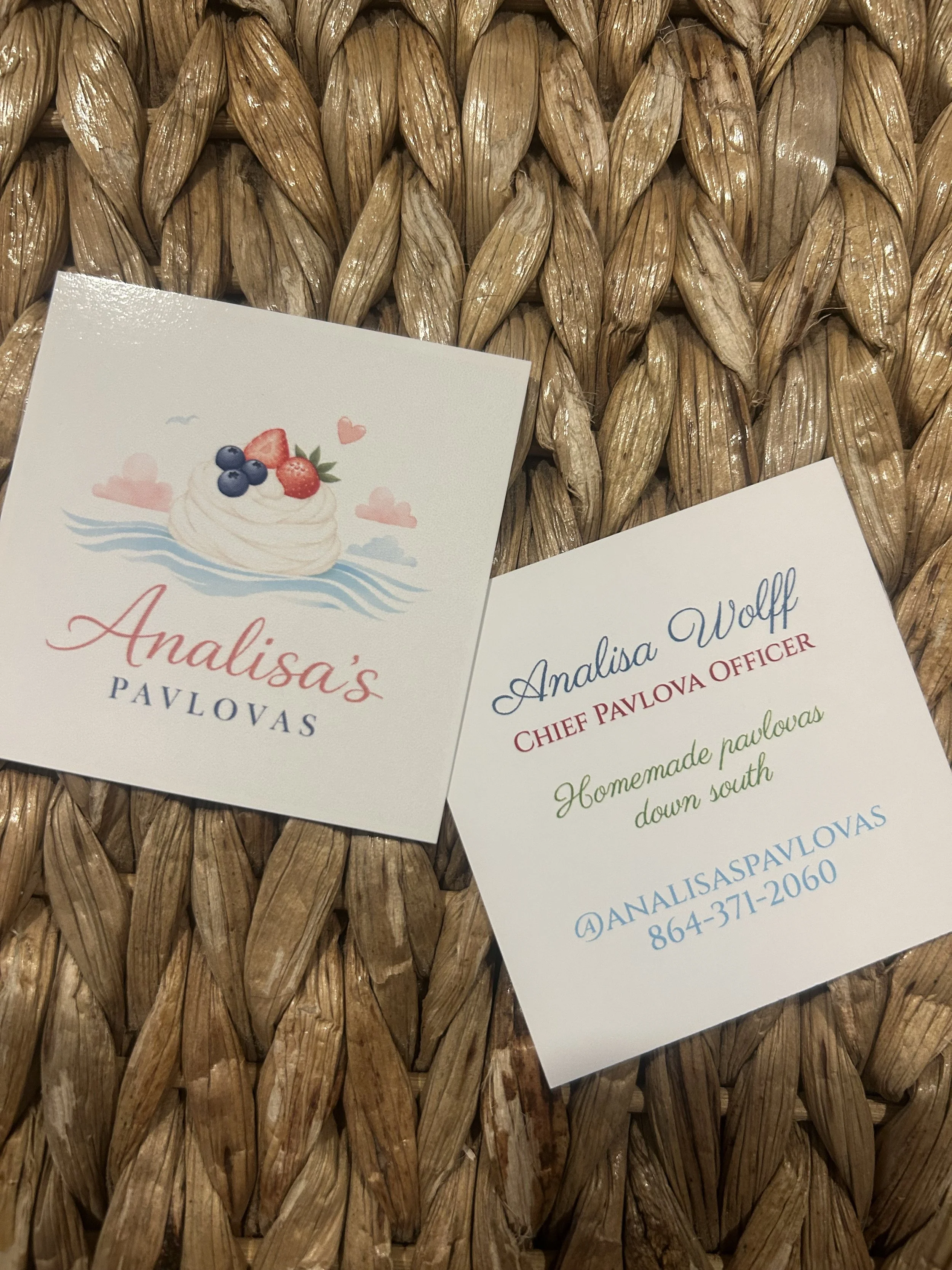 Close-up of a woven straw surface with business cards on top, one featuring a dessert with berries illustration and the other with text and contact information.