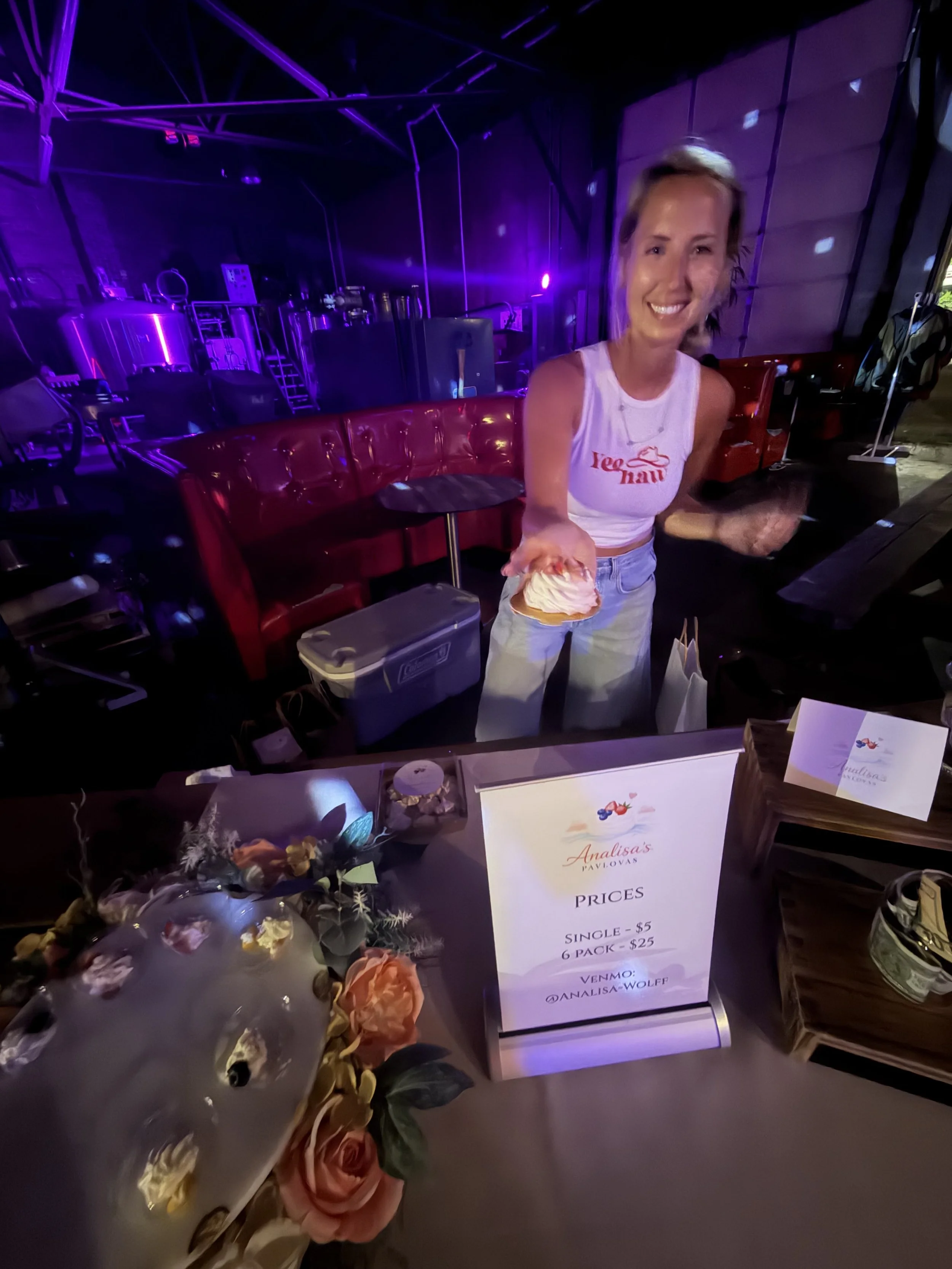 A woman handing an ice cream cone to the camera at an indoor event with purple lighting. There are flowers and a sign listing prices for ice cream on the table.