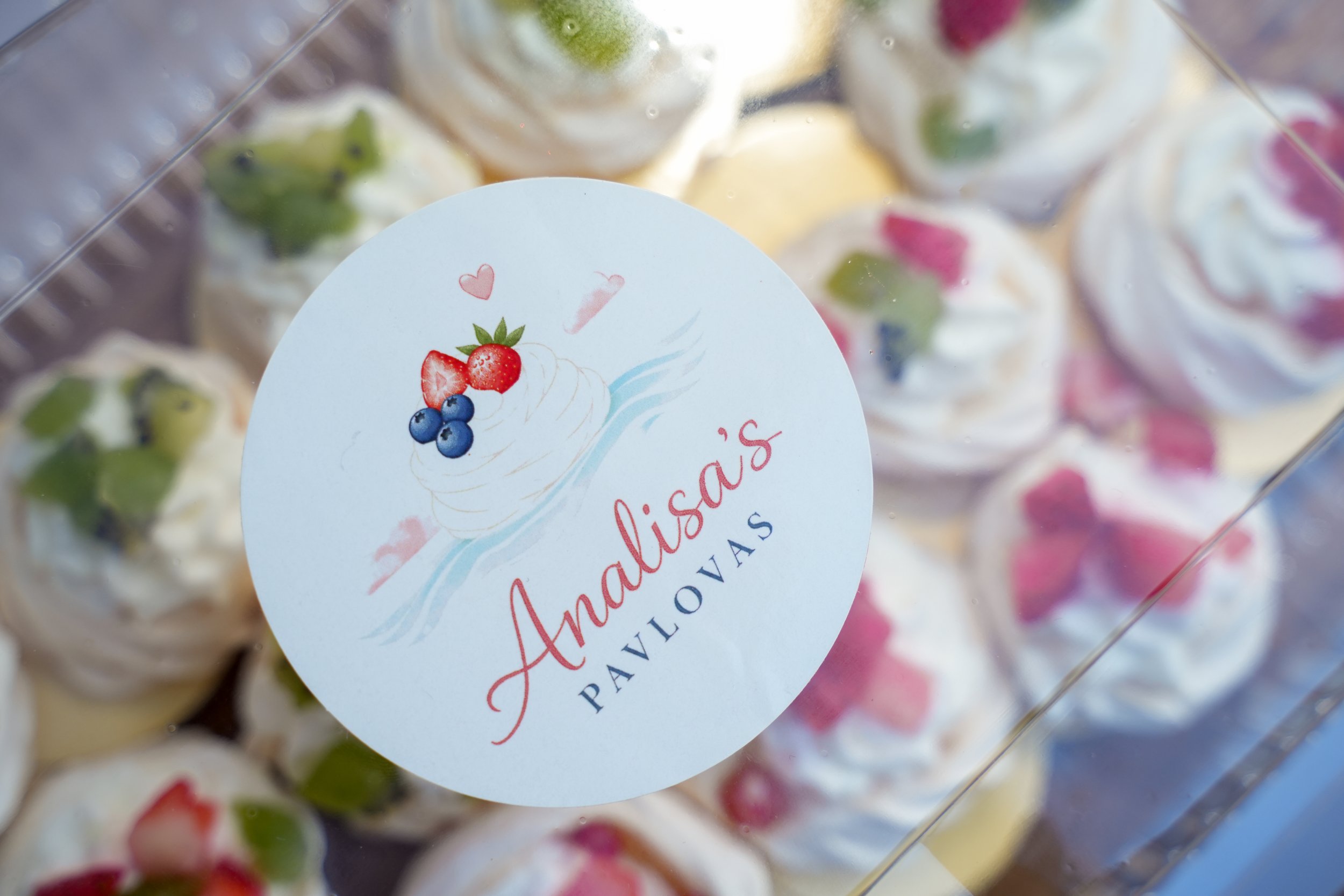 Close-up of a clear container filled with assorted fruit-flavored ice creams or gelato with colorful fruit pieces. A round white label with a printed illustration of a scoop of ice cream topped with strawberries, blueberries, and hearts is attached to the container.