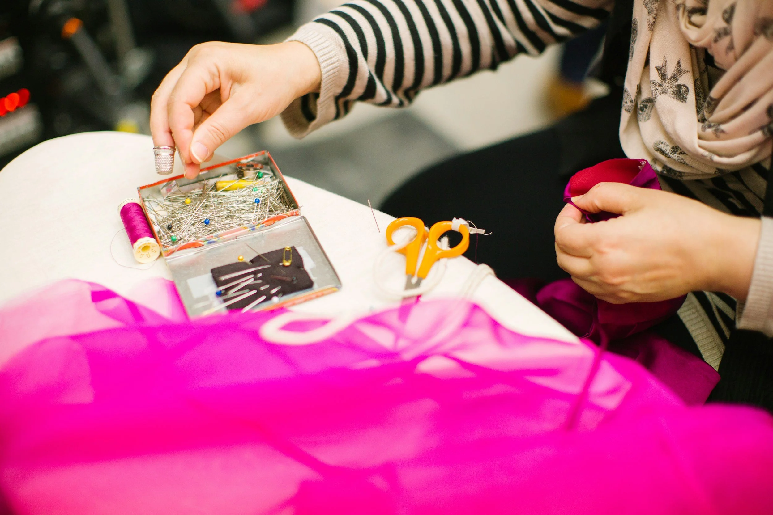 Close-up of a person sewing or doing needlework with a box of assorted sewing pins, a pink thread spool, a pair of scissors, and pink fabric on a table.
