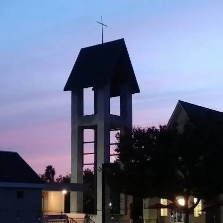 Church steeple with cross at sunset, surrounding houses and trees.