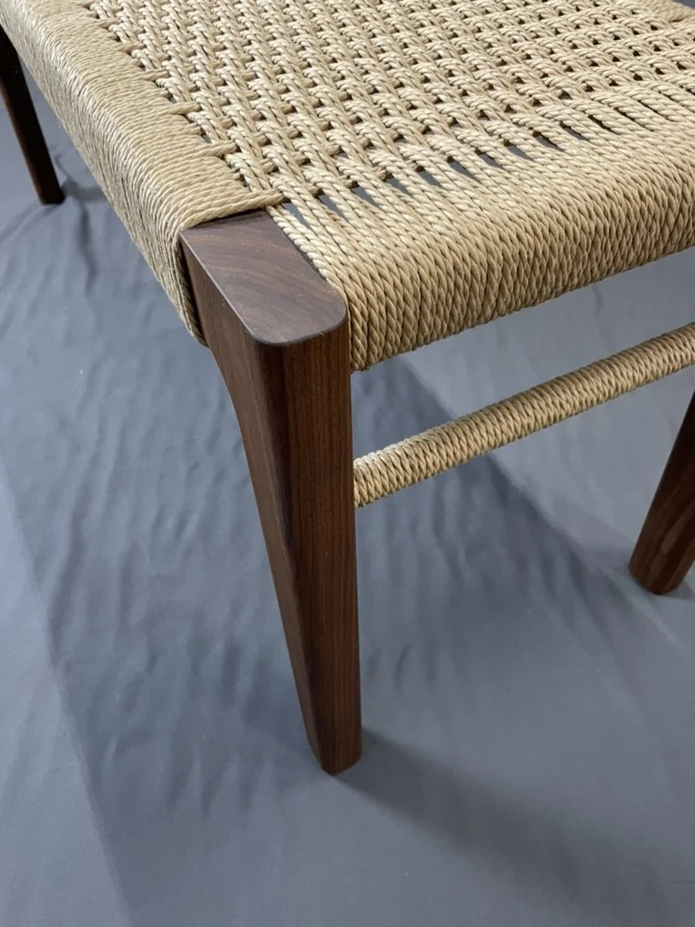 Close-up of a wooden bench leg made with solid walnut with a Danish cord woven seat, against a gray floor.