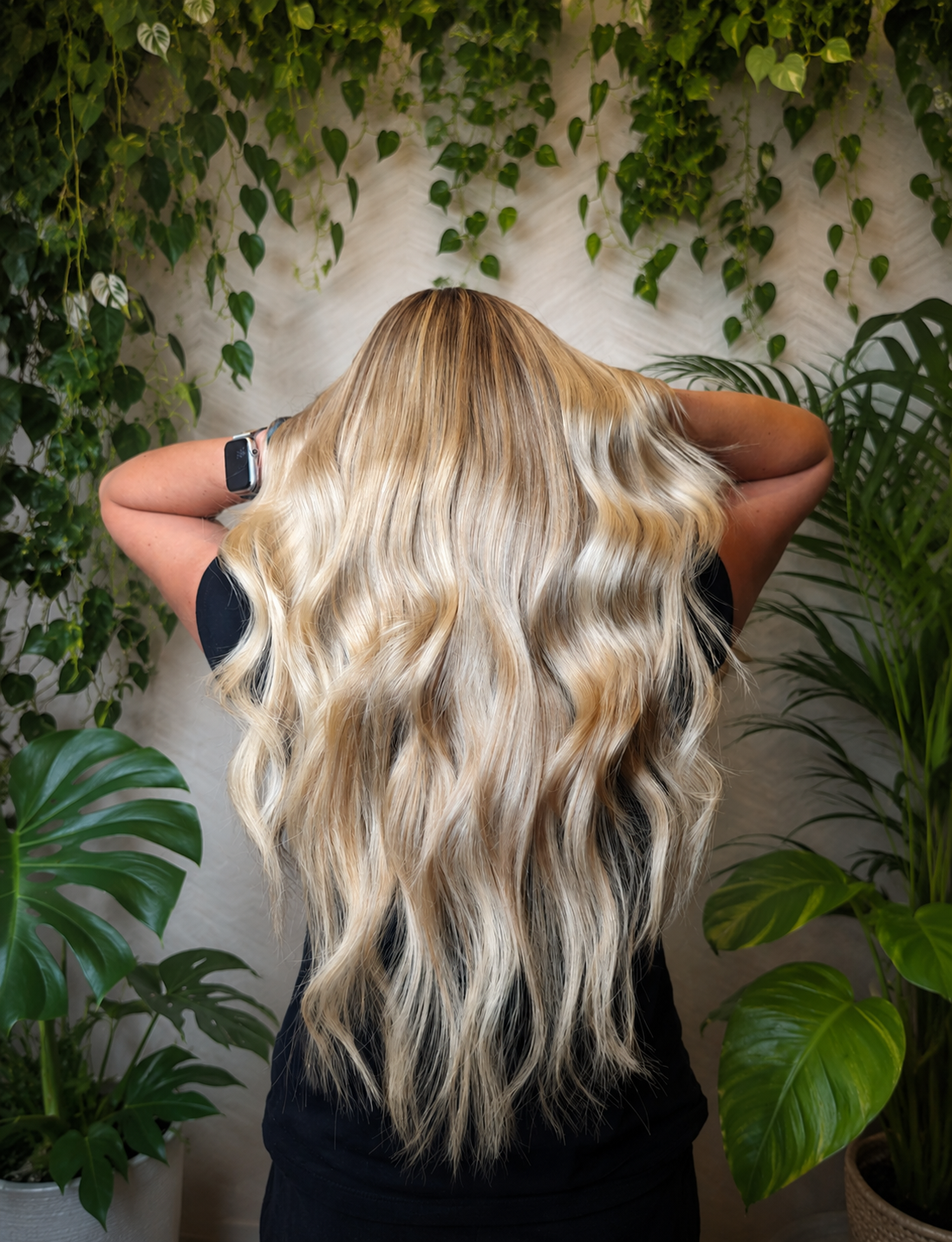 Back of a person with long, wavy blonde hair standing in front of a wall with green plants.