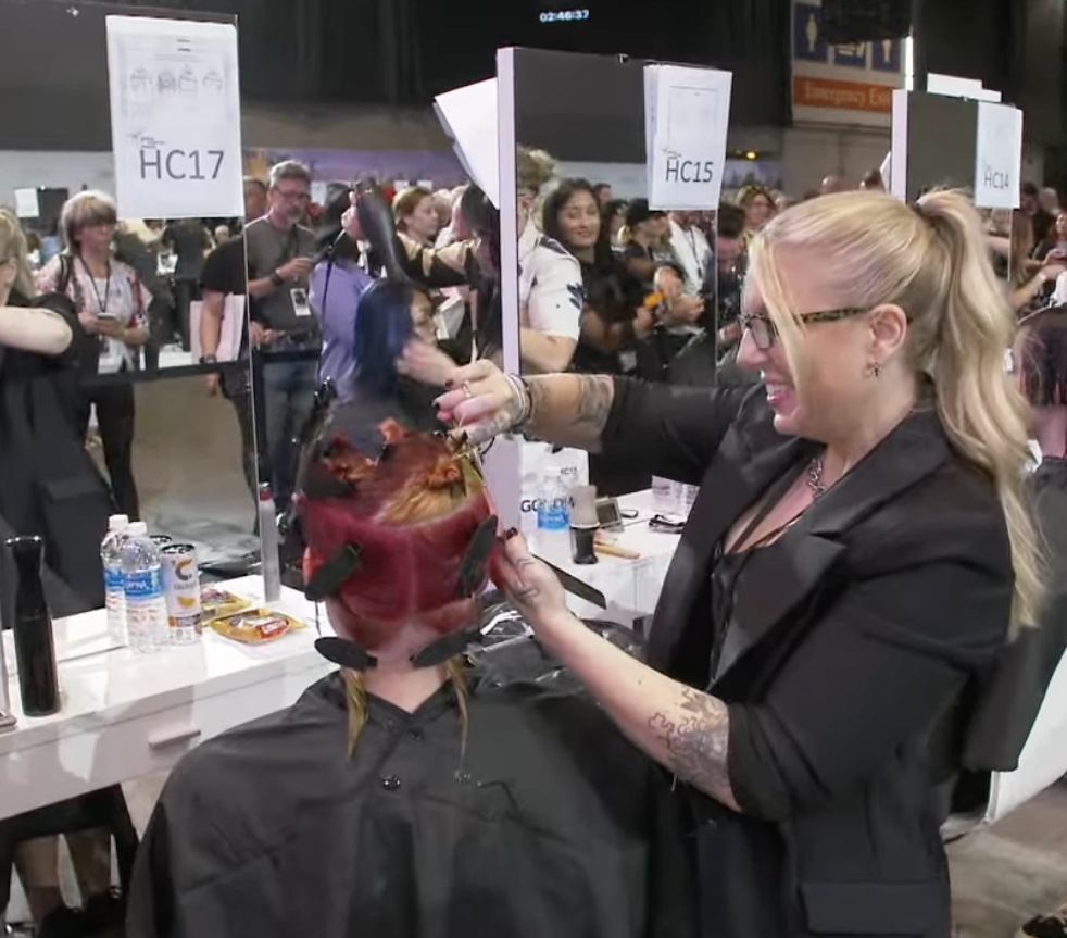 A woman with glasses and a high ponytail applies hair color to a person's hair at a busy hair color demonstration event.