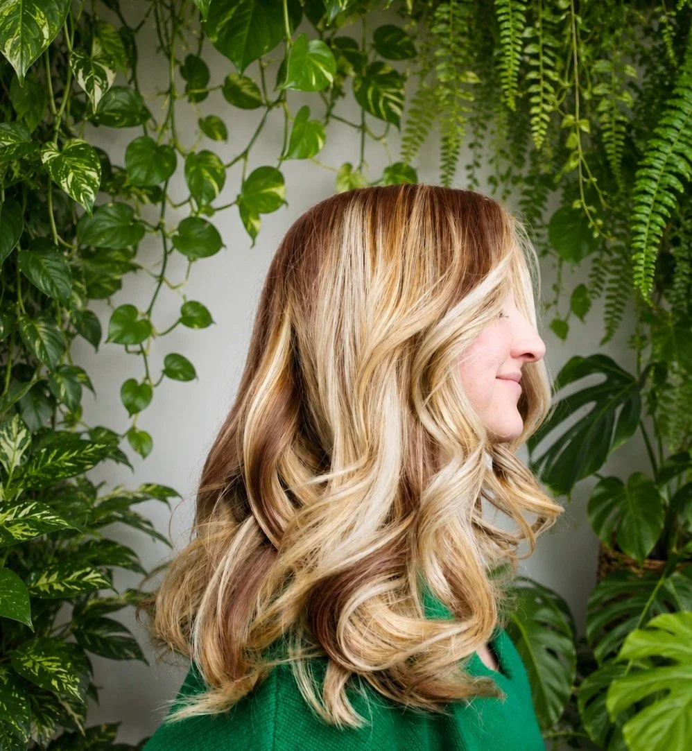 A woman with blonde, wavy hair standing among green plants with large leaves and fern-like fronds.