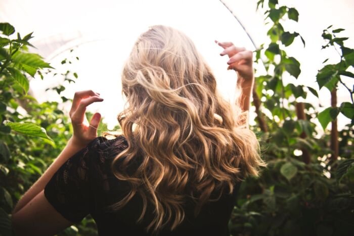 Back view of a woman with curly blonde hair standing amidst green plants inside a greenhouse or garden with natural light.