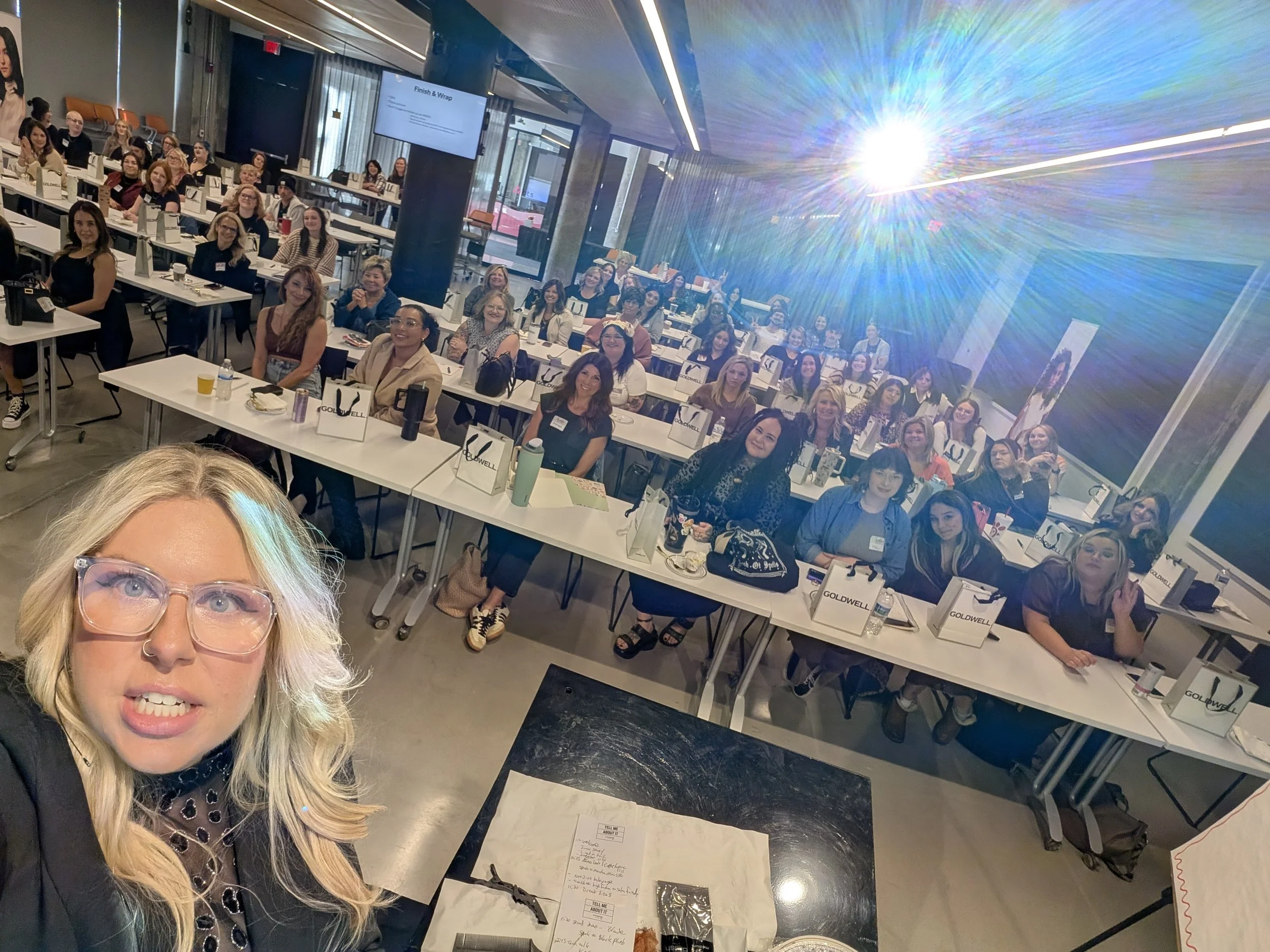 A woman with blonde hair, glasses, and a nose ring taking a selfie in front of a large group of women seated at tables in a conference room. The women are smiling and facing the camera, with some holding beverages and others wearing name tags. A presentation slide on the screen behind them reads 'Finish & Wrap'.