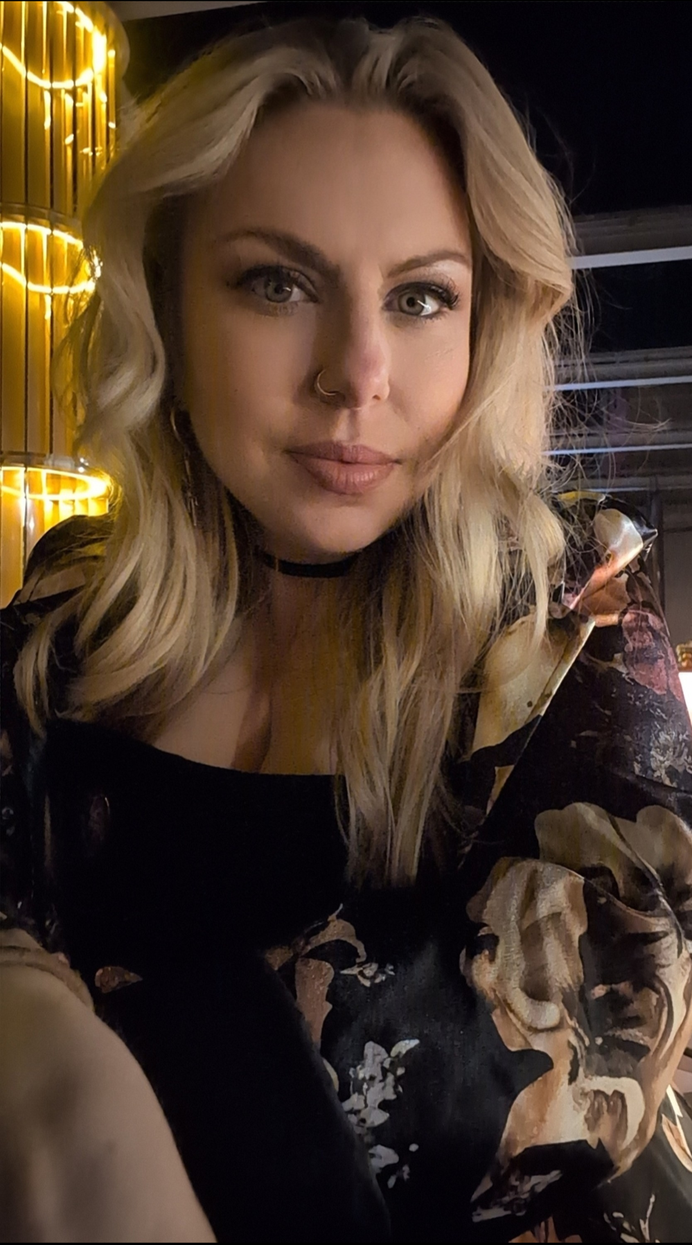A woman with blonde wavy hair, wearing a black choker, lipstick, and eye makeup, taking a selfie in a dimly lit environment with warm lighting and a dark background.