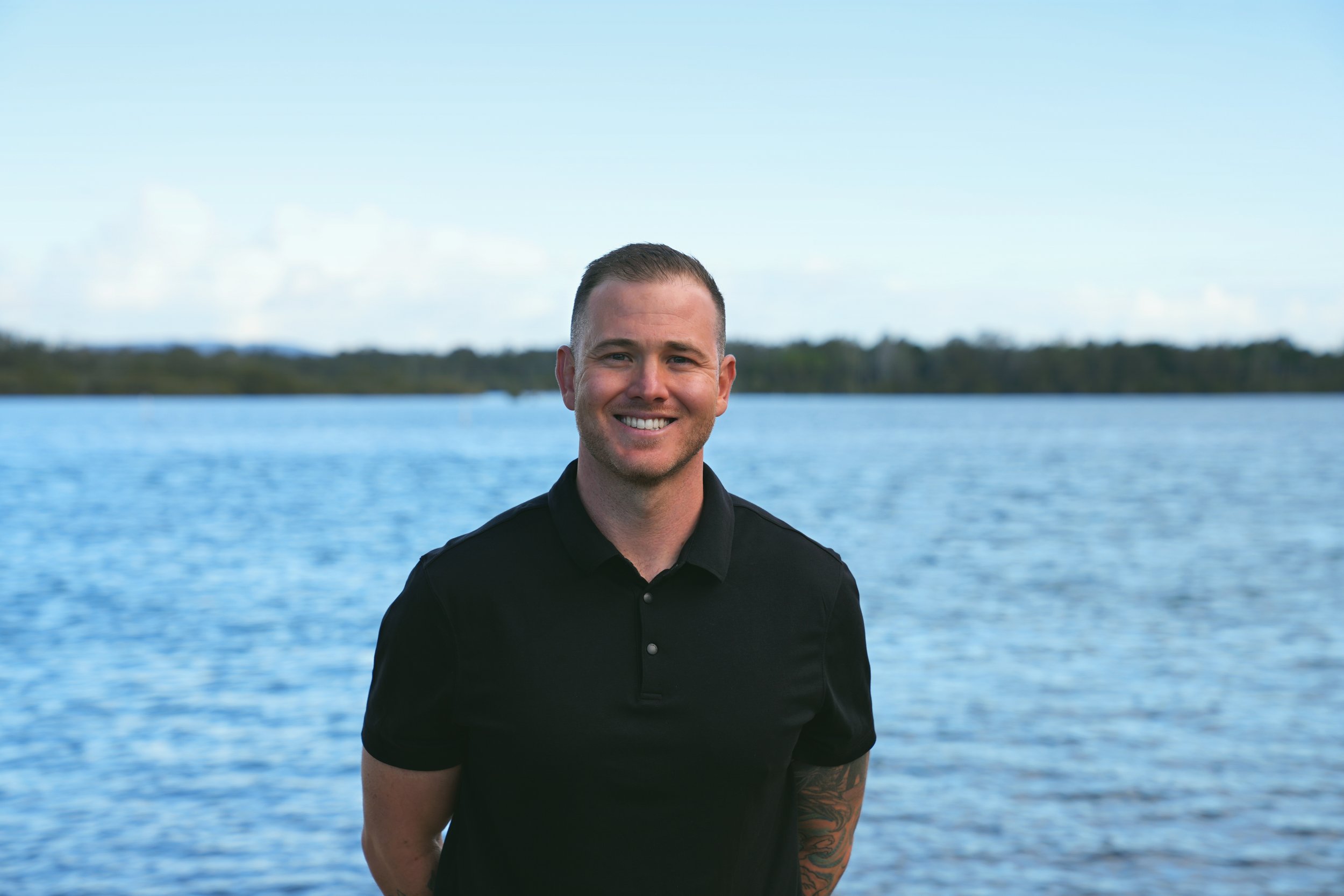 A smiling young man with short brown hair, wearing a black shirt, stands outdoors near a body of water with an island or shoreline in the background under a partly cloudy sky.