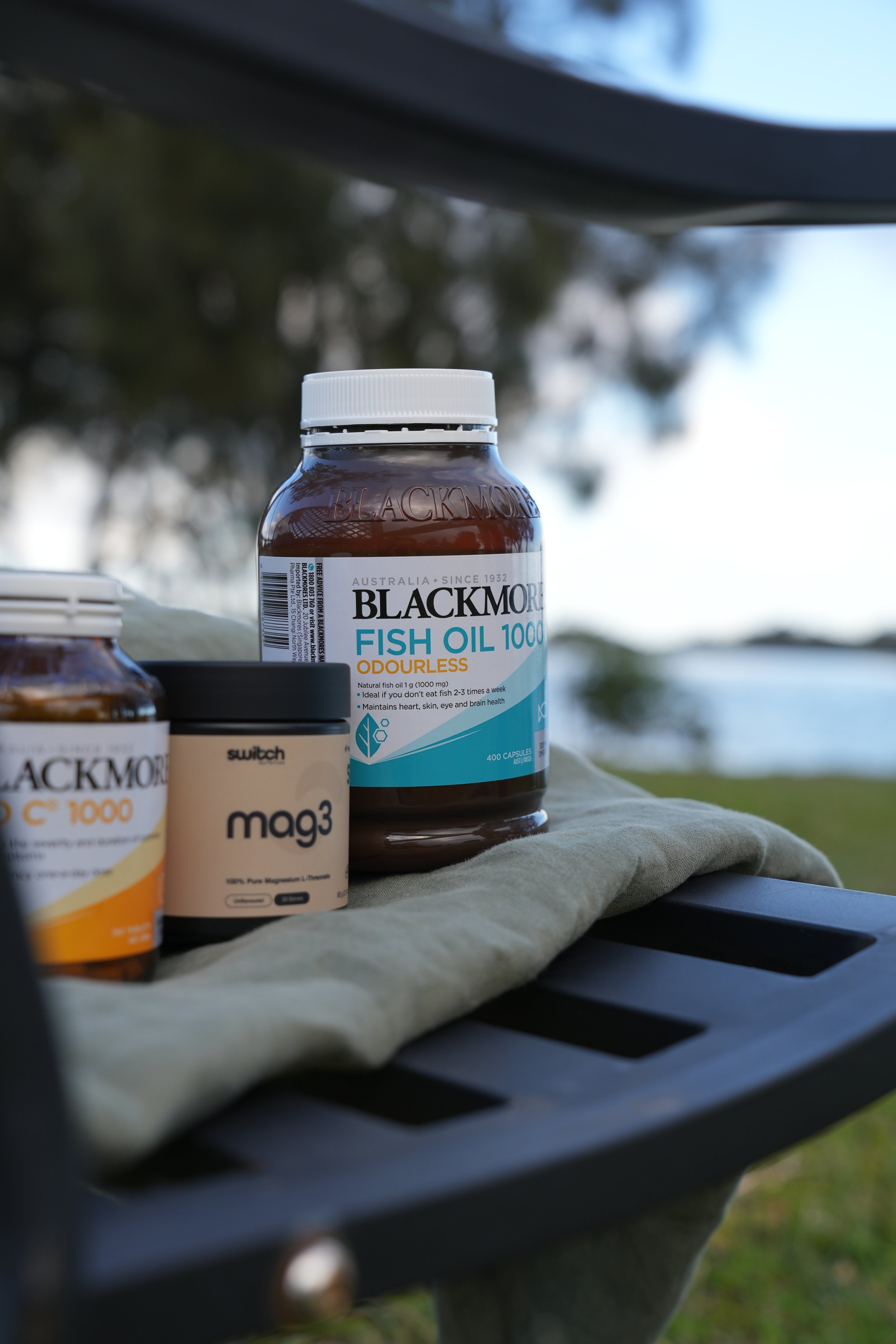Close-up of dietary supplements on a black surface with blurred outdoor background. Visible are bottles of Blackmores Fish Oil 1000, Mag3, and Switch.