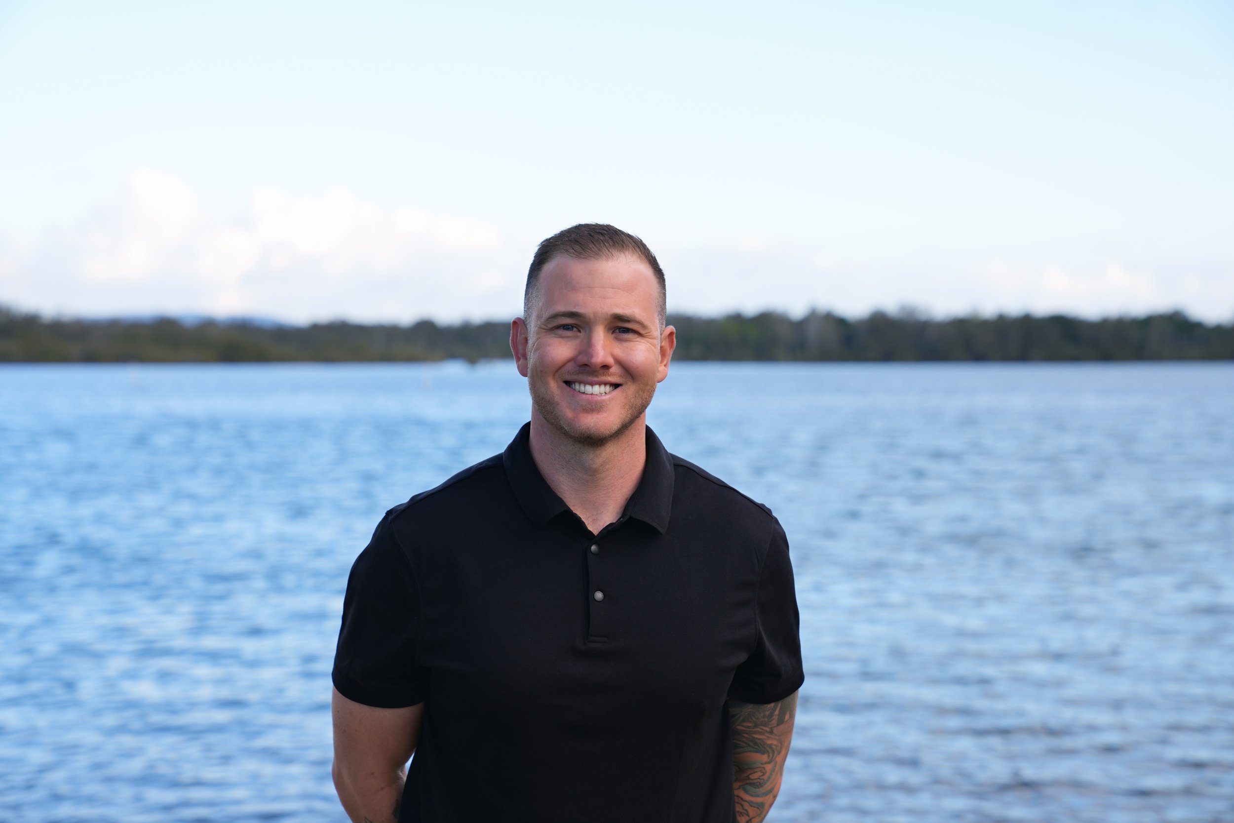 A smiling man standing near a body of water with a distant shoreline and trees, wearing a black shirt.